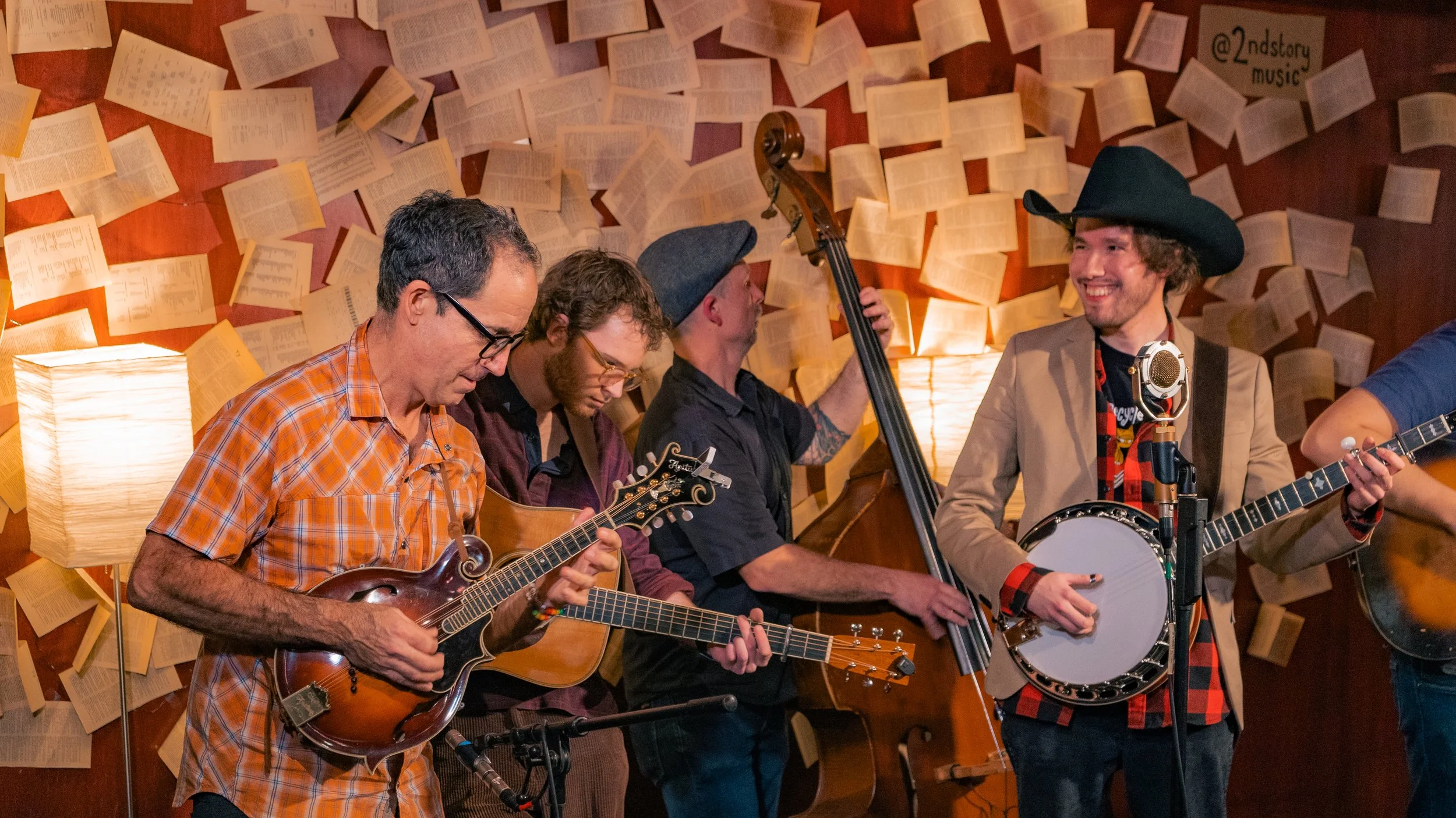 Group of five musicians playing instruments on stage with pages of sheet music on the wall behind them, warm lighting, and a vintage microphone.