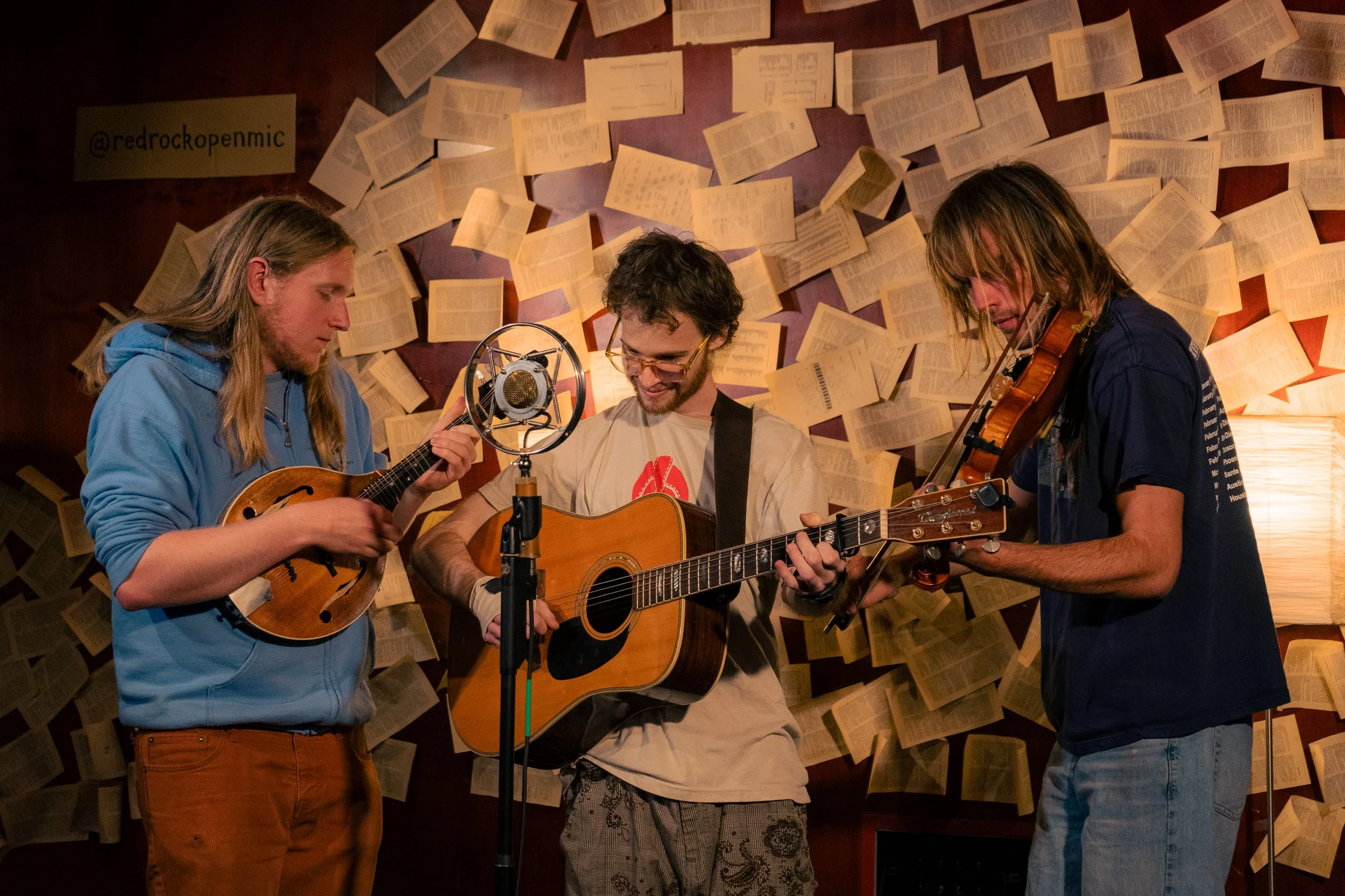 Three young men playing musical instruments in front of a wall decorated with open books, with a vintage microphone in the center.