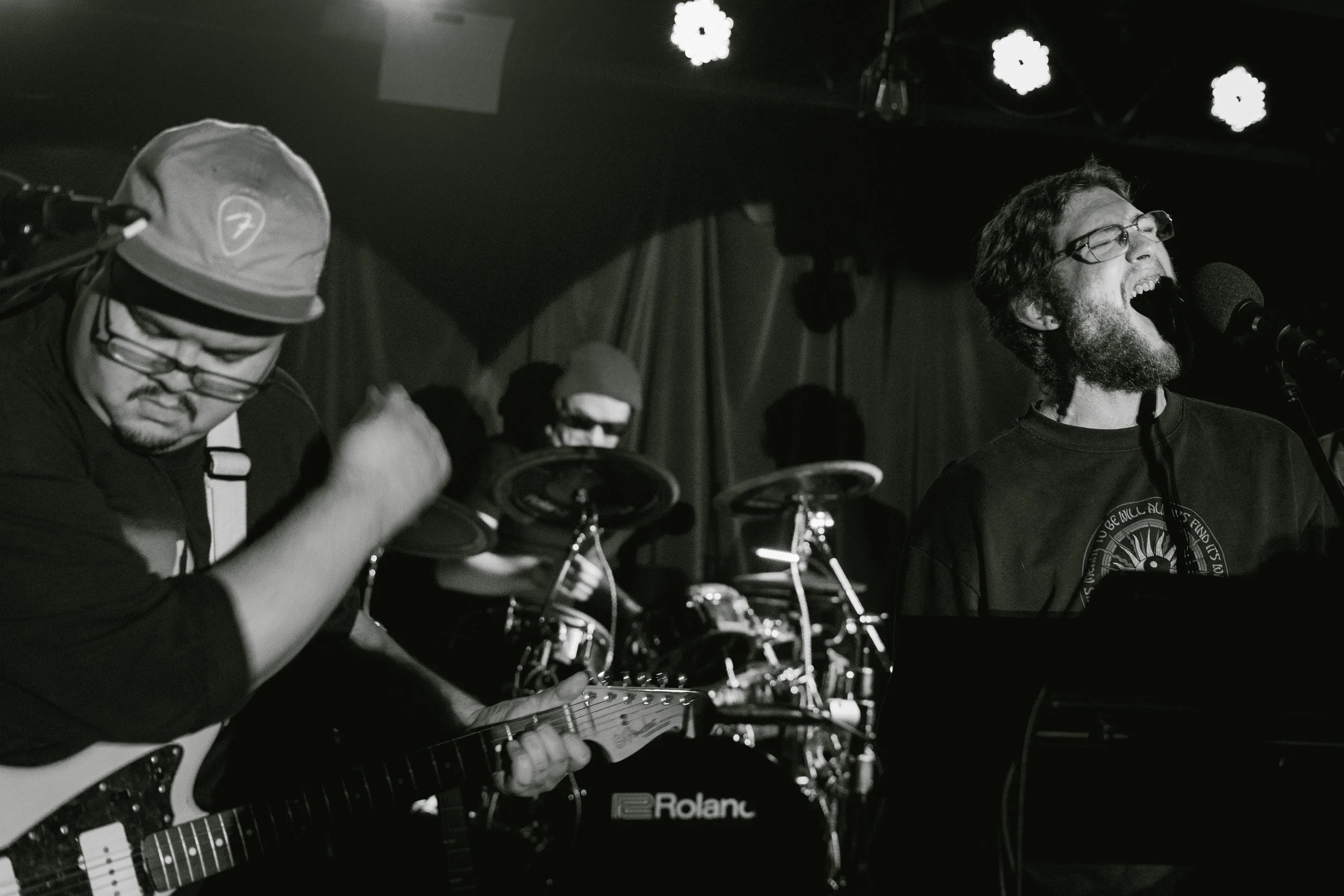 Black and white photo of a band performing live on stage, including a guitar player wearing glasses and a beanie, a drummer wearing sunglasses and a beanie, and a vocalist singing into a microphone with glasses and a beard.