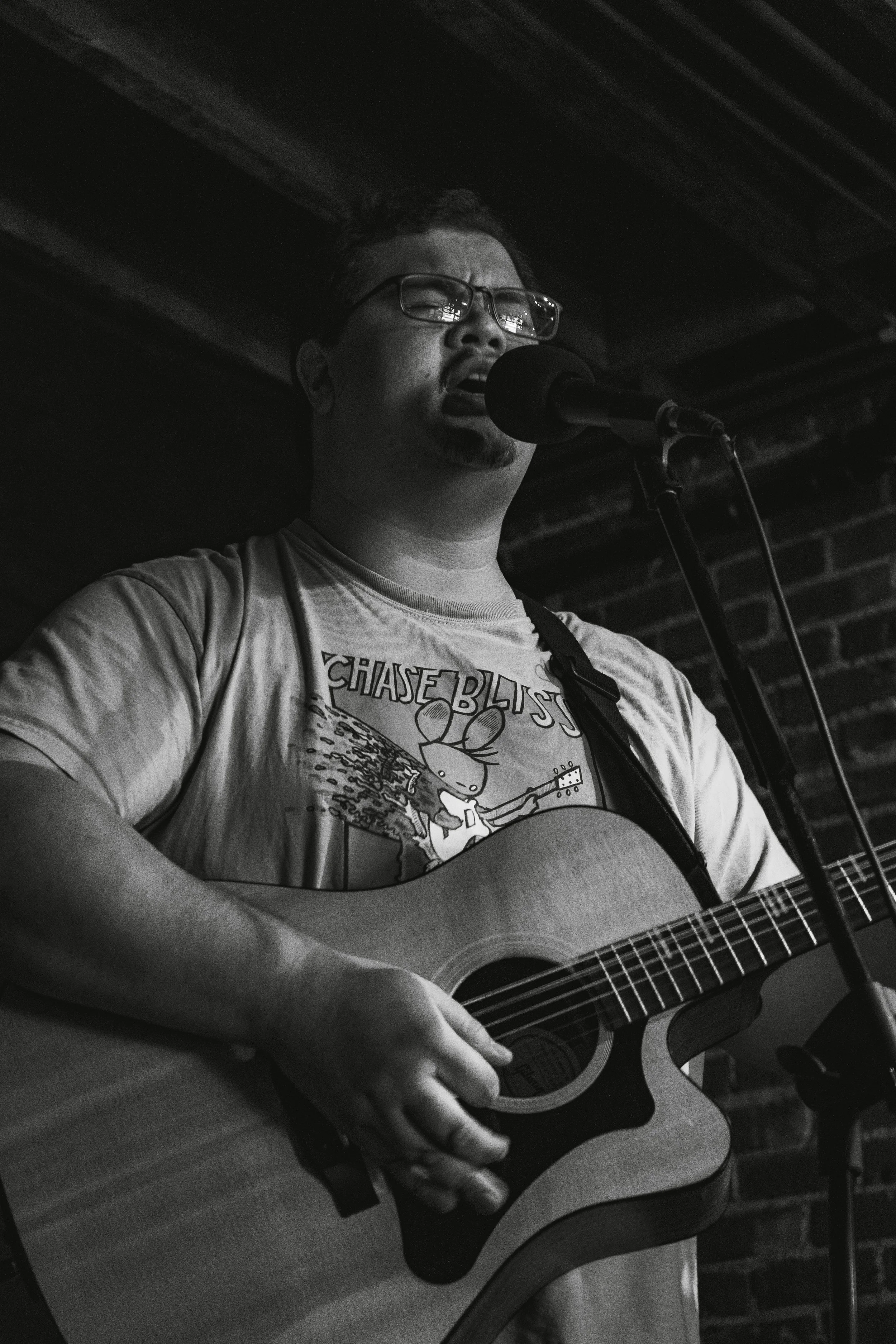 A man with glasses singing into a microphone while playing an acoustic guitar in a dimly lit venue with exposed brick wall.