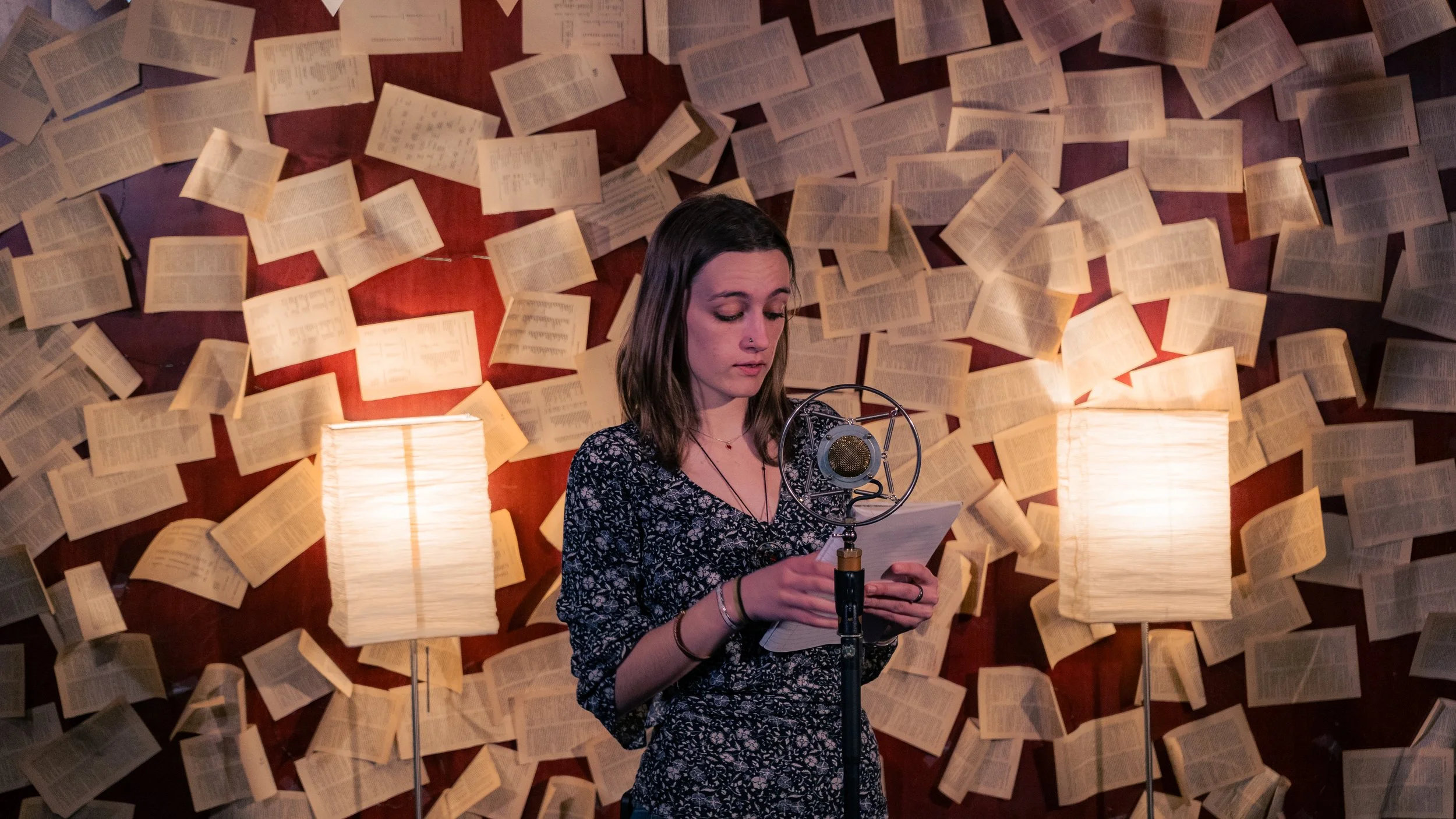 A young woman standing in front of a wall decorated with open books, reading into a microphone while holding papers, with two lamps illuminating the scene.