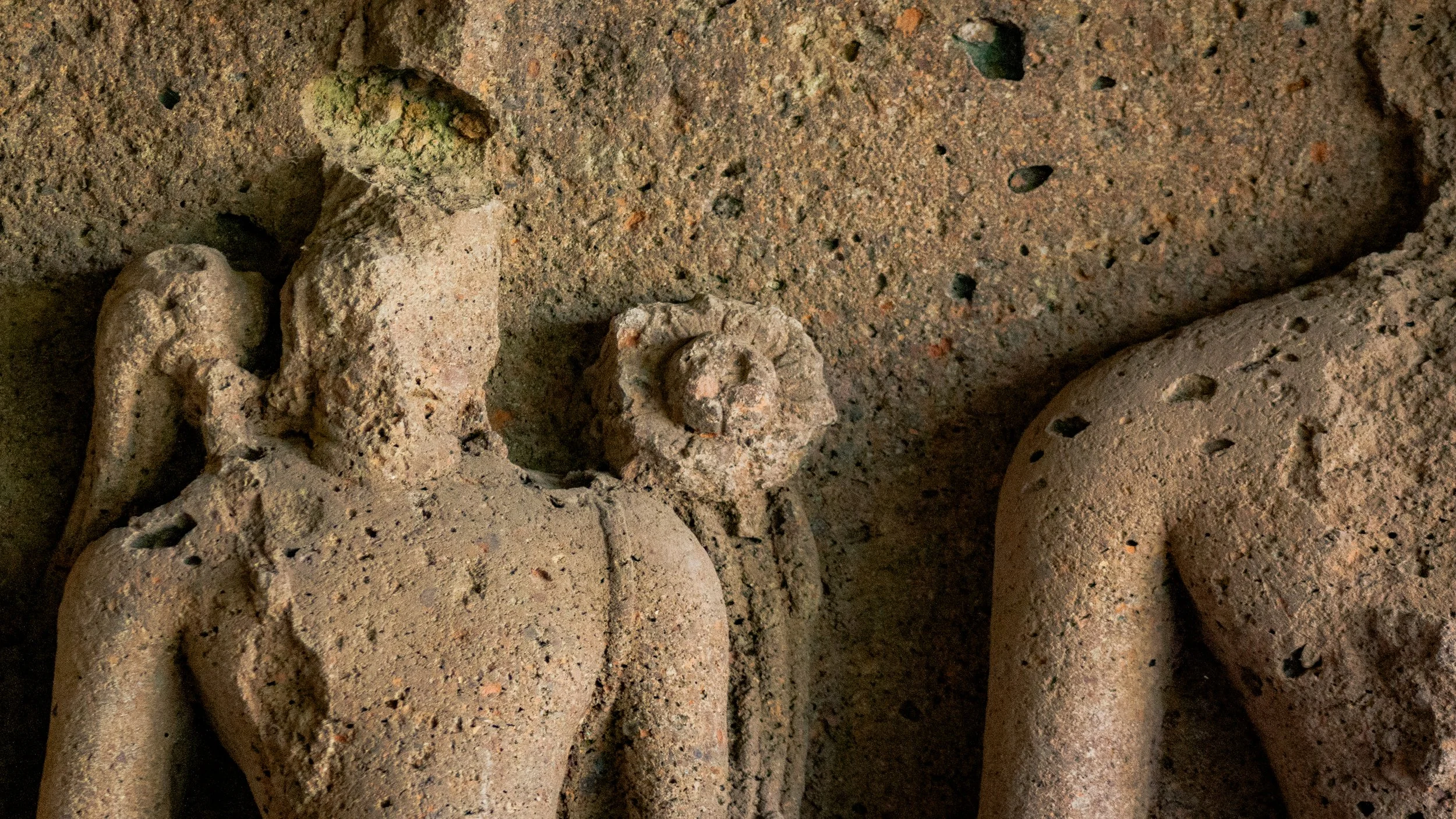 Close-up image of an ancient clay sculpture of a humanoid figure with a coiled object on its side, and part of another figure visible on the right.