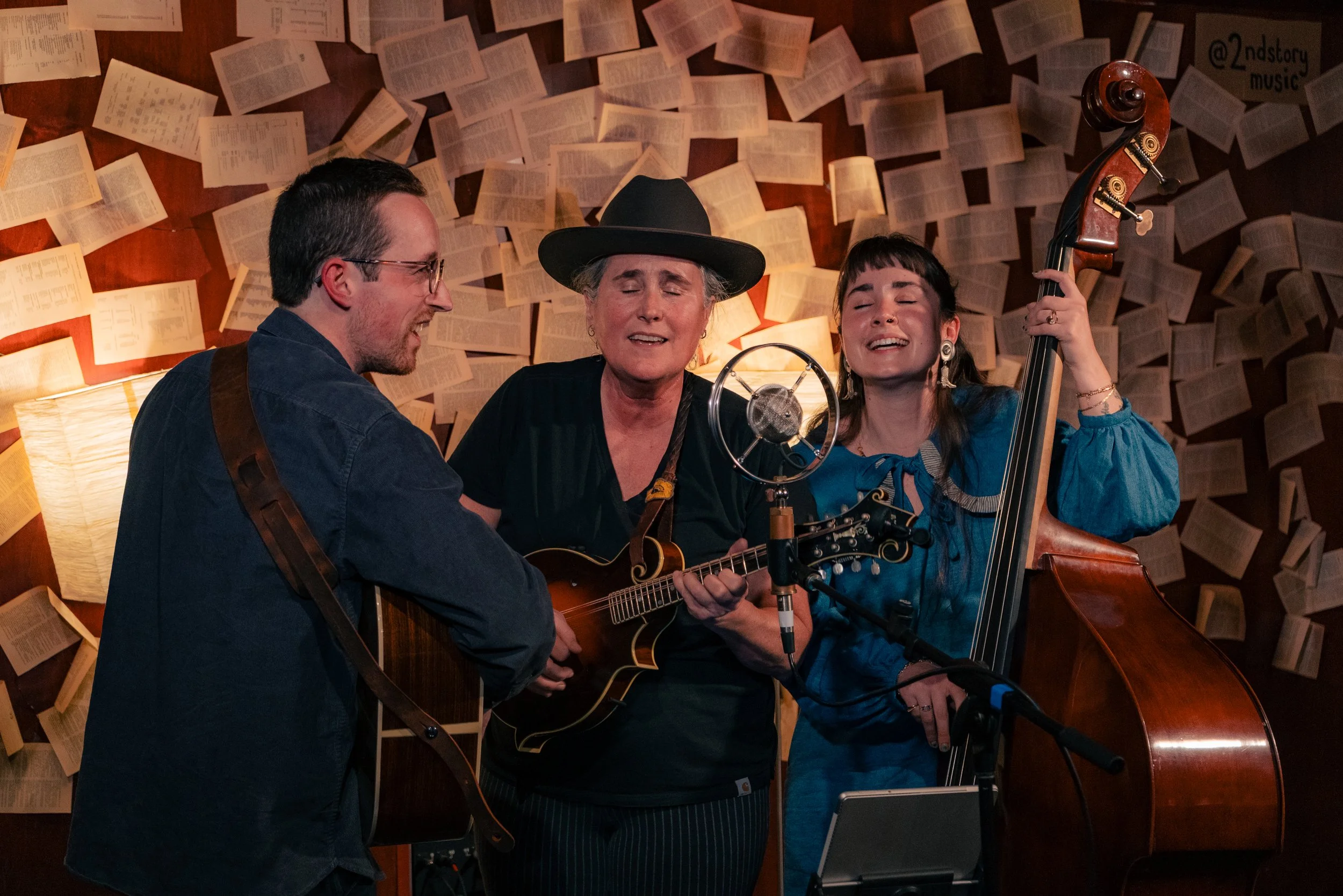 Three musicians singing and playing instruments in a cozy room with walls covered in open books, with a microphone in front of them.
