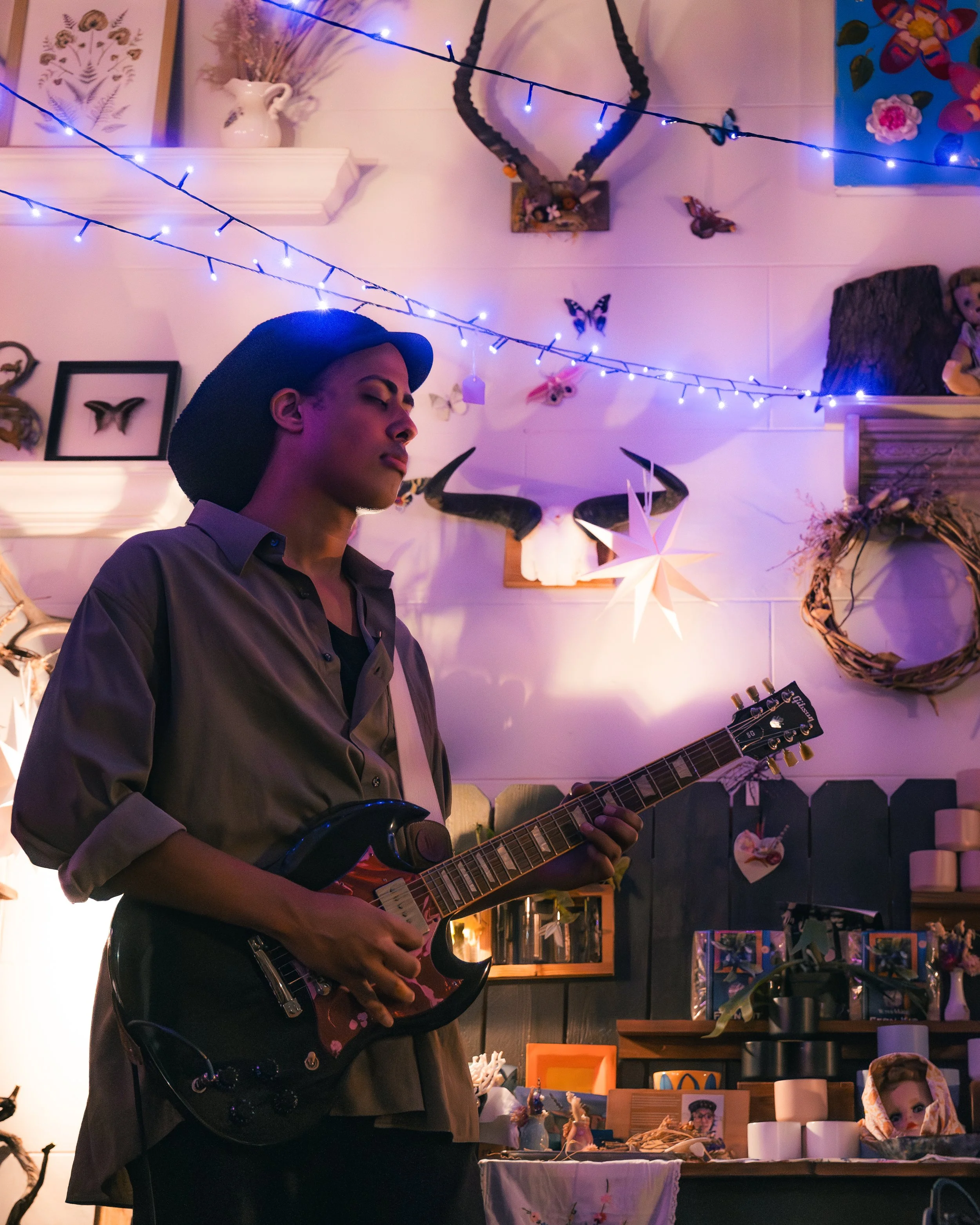 A young person playing a black electric guitar in a room decorated with string lights, butterflies, a star-shaped paper lantern, and various decorative items and artwork.
