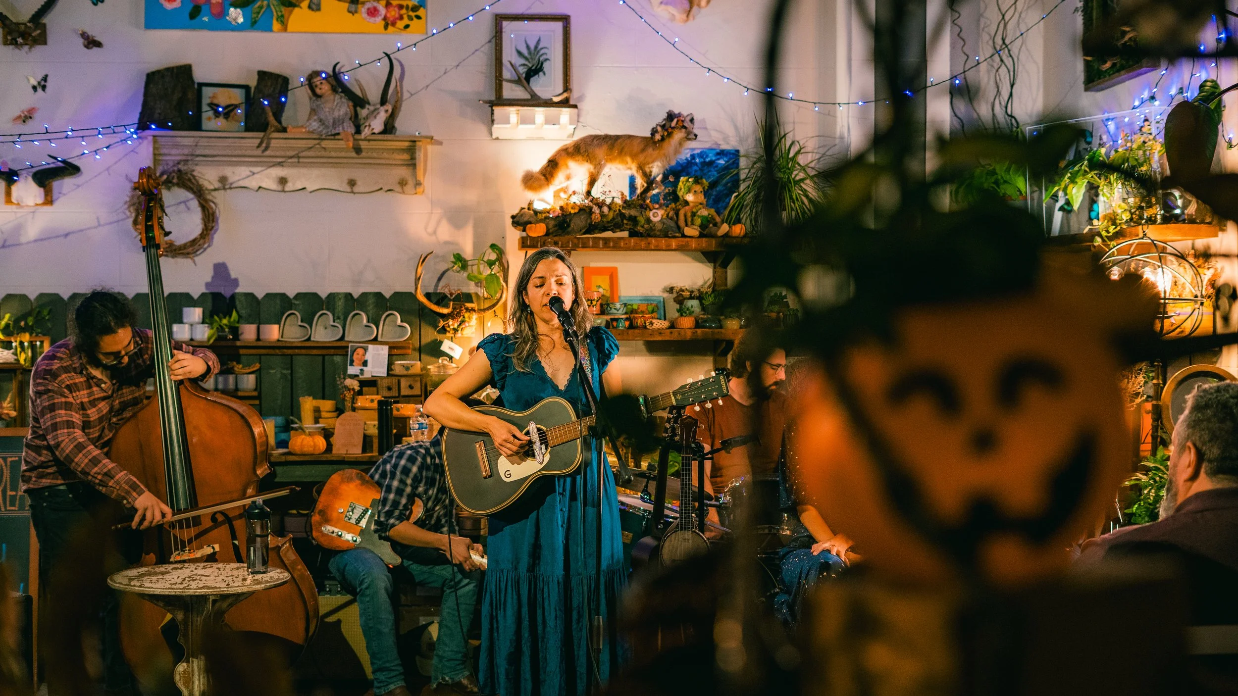 A musical performance in a cozy, decorated venue with a woman singing and playing guitar, a man playing double bass, and another musician in the background. The scene includes Halloween decorations and warm lighting.
