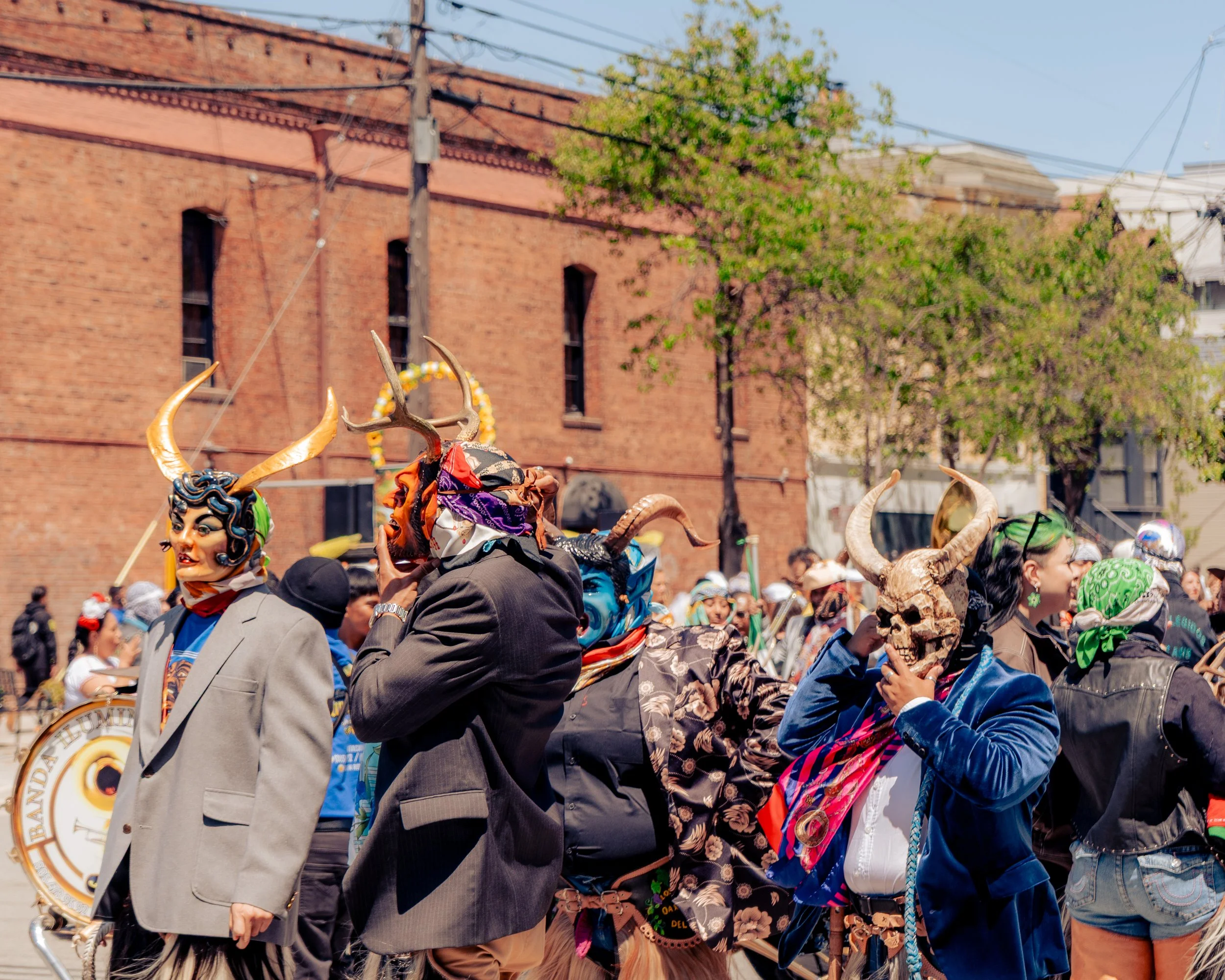 People wearing colorful costumes and masks with horns and skulls, participating in a street parade in front of a red brick building.