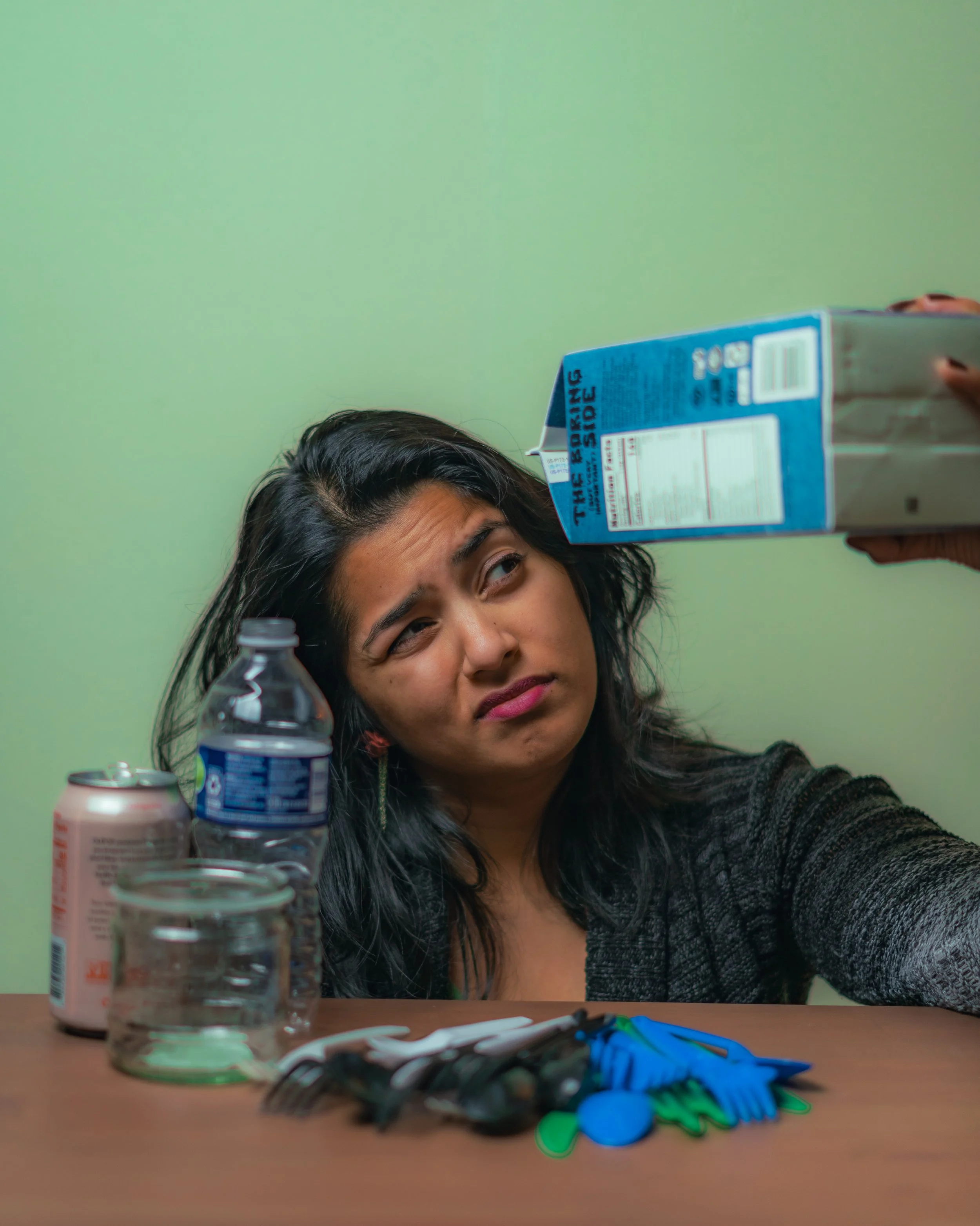 A woman sitting at a table looks annoyed as another person holds a bag of snacks above her head. The table has a water bottle, a milk canister, a glass, some utensils, and small toy spoons and forks.
