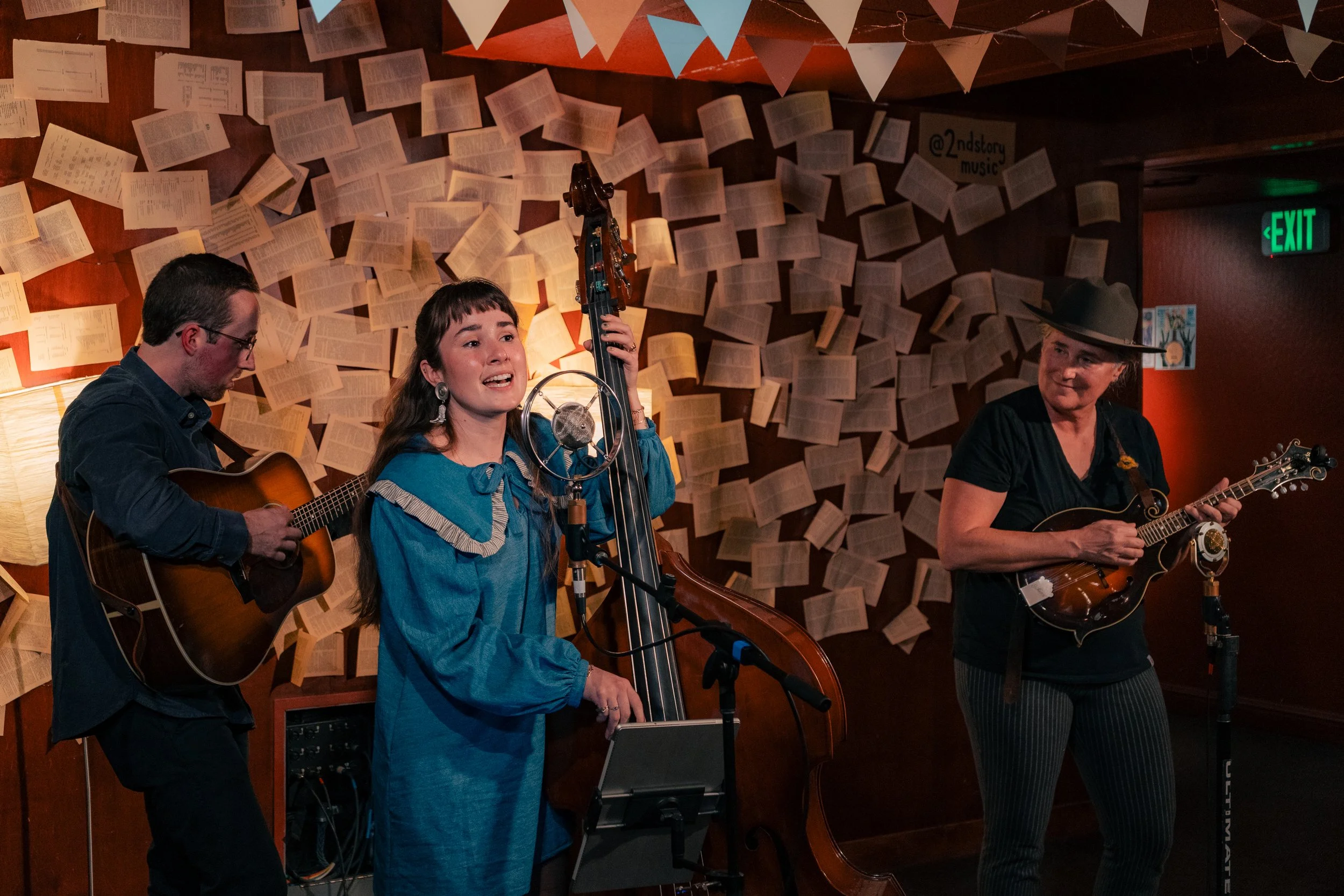 Three musicians perform in front of a wall covered with open books, with colorful pennant banners hanging above them. One plays guitar, the woman sings into a microphone while playing a double bass, and another woman plays mandolin.