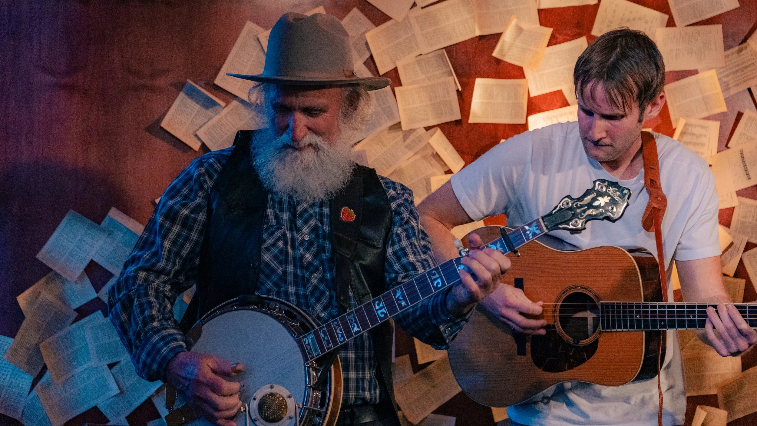 Two men playing guitars, one with a banjo, in front of a wall covered with open books or pages.