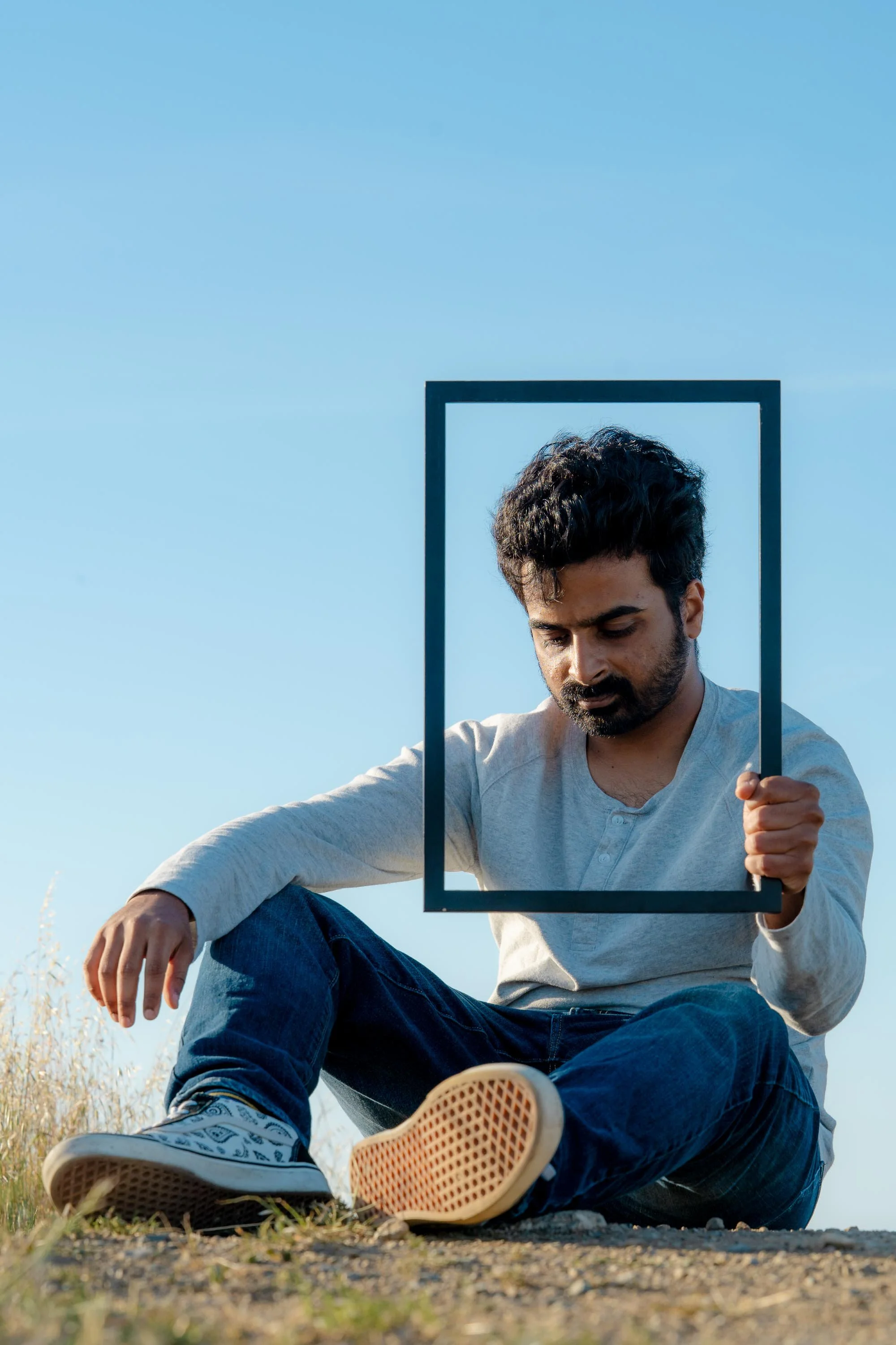 A man sitting outdoors on a ground with grass, holding a black rectangular frame, with a clear blue sky in the background.