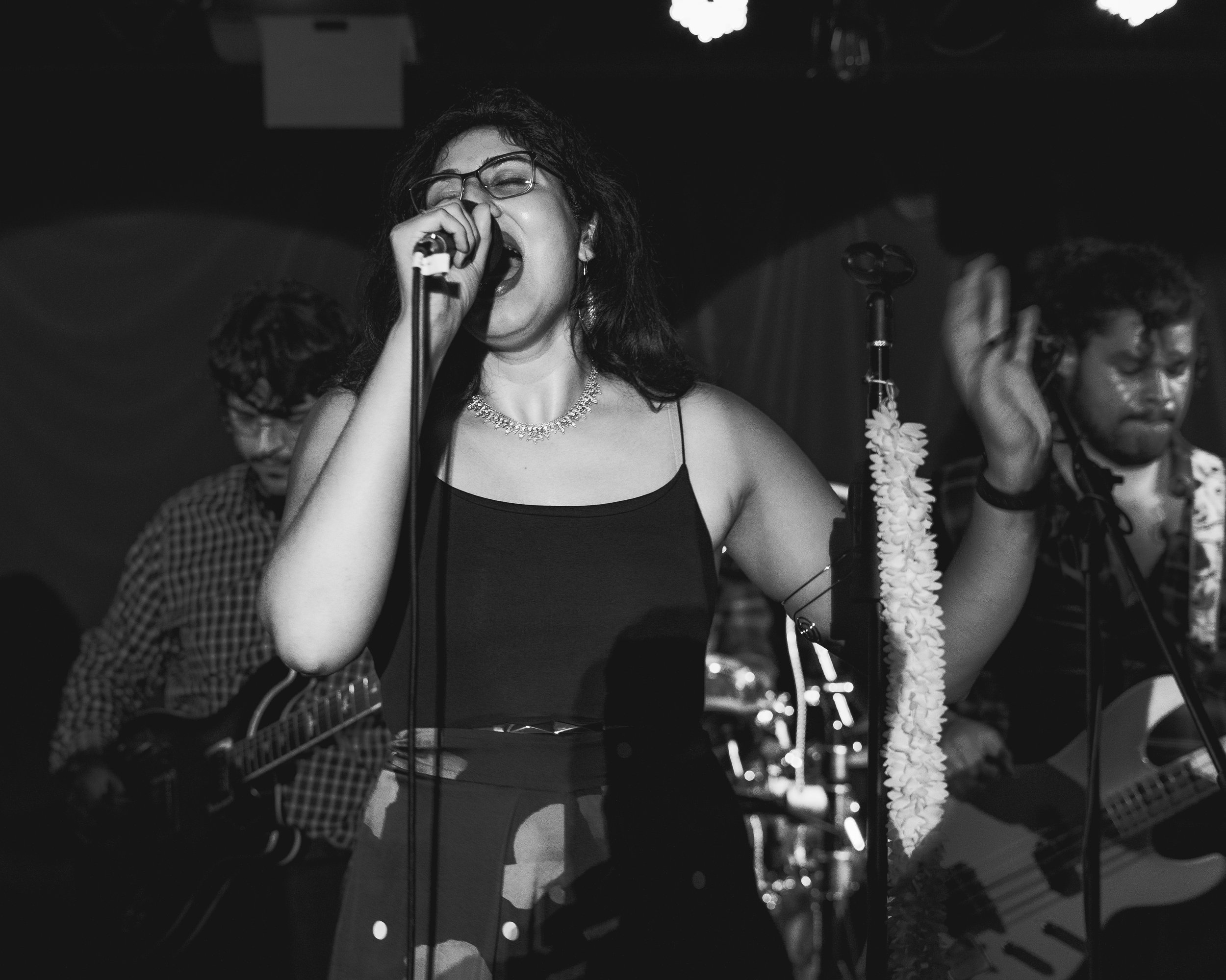 A woman singing passionately into a microphone during a live music performance, with two musicians playing guitars beside her, in a dimly lit venue.