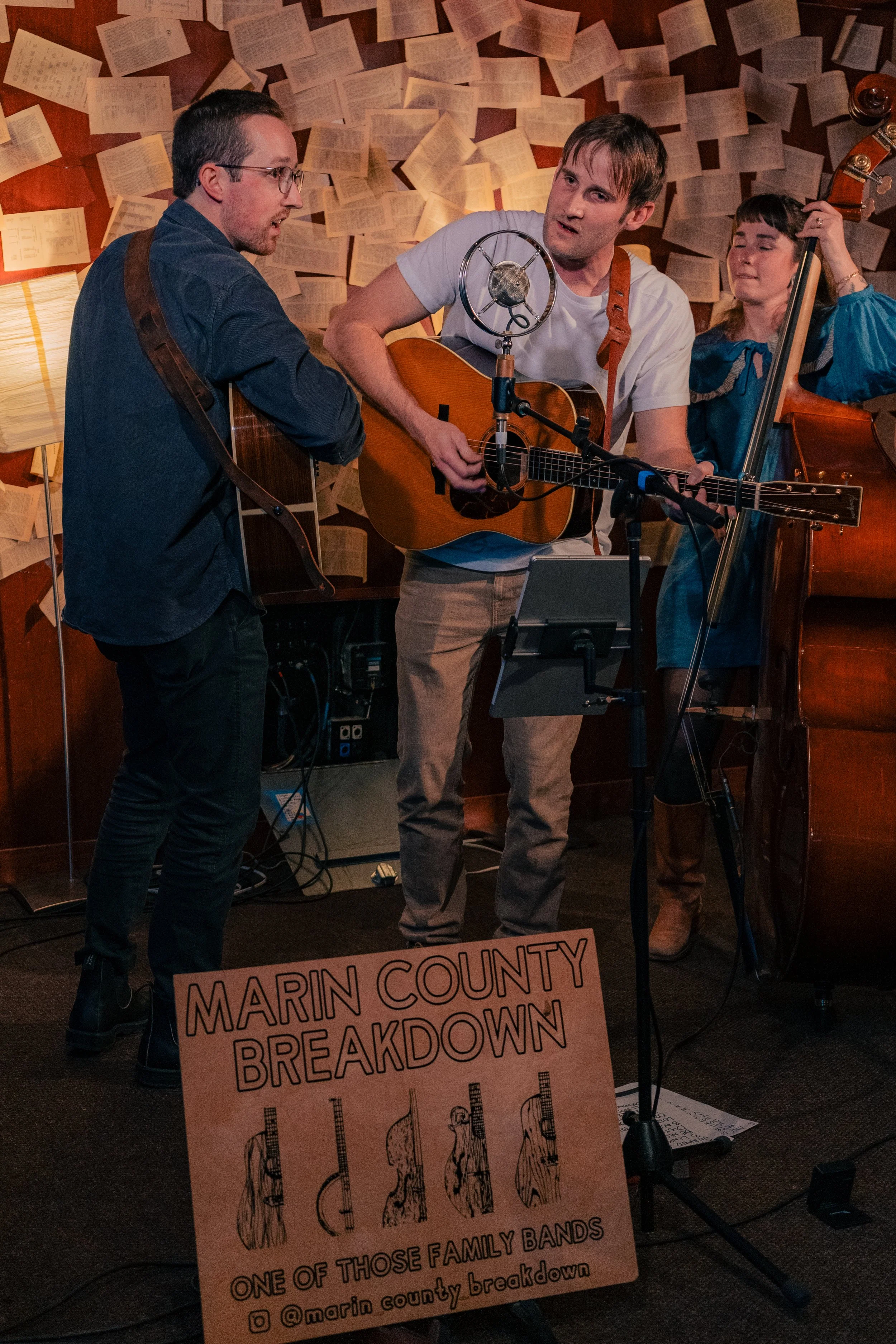 Three musicians performing live, with two men playing guitars and a woman playing a double bass, in a room decorated with open books on the walls. A sign on the floor reads 'Marin County Breakdown, One of Those Family Bands, @marin_county_breakdown'.