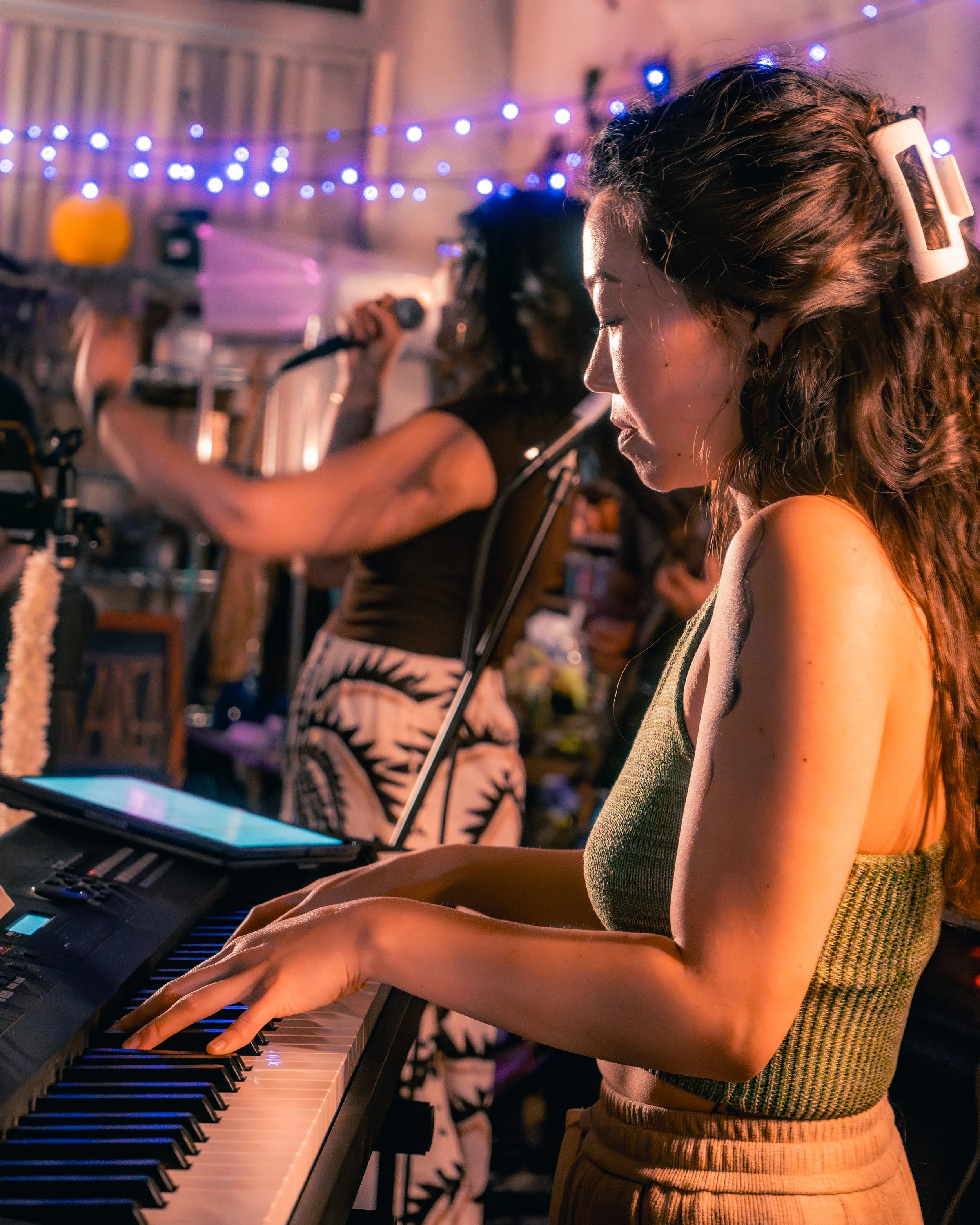 Woman playing keyboard indoors during a music session with string lights overhead and another singer in the background.
