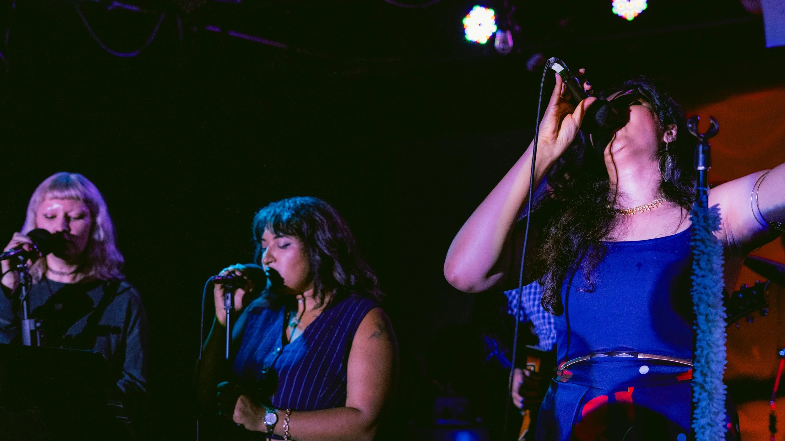 Three women singing into microphones on stage, with colorful stage lights overhead.