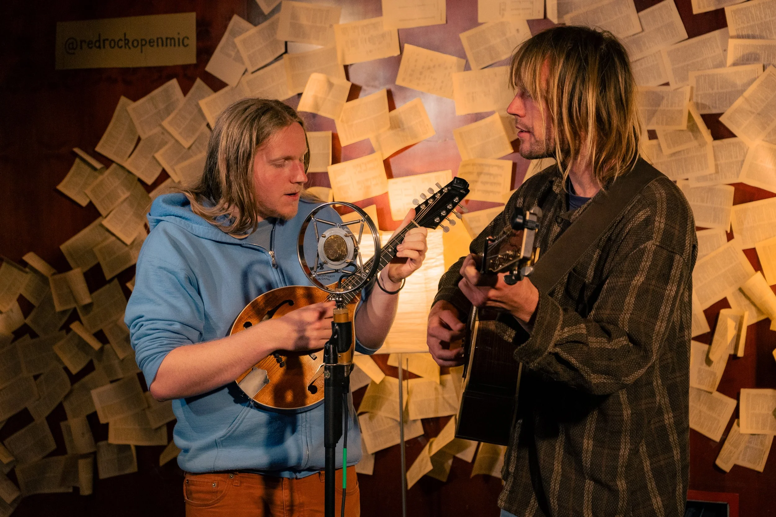 Two men playing acoustic guitars and singing in a cozy room decorated with open books on the wall. One man in a blue hoodie is singing into a vintage microphone, while the other in a dark plaid shirt is looking at him, playing his guitar.