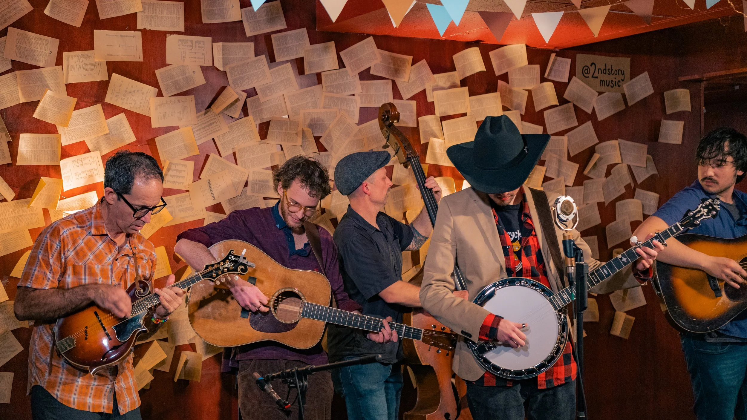 Five musicians playing instruments in a room with walls decorated with open books and a colorful bunting.