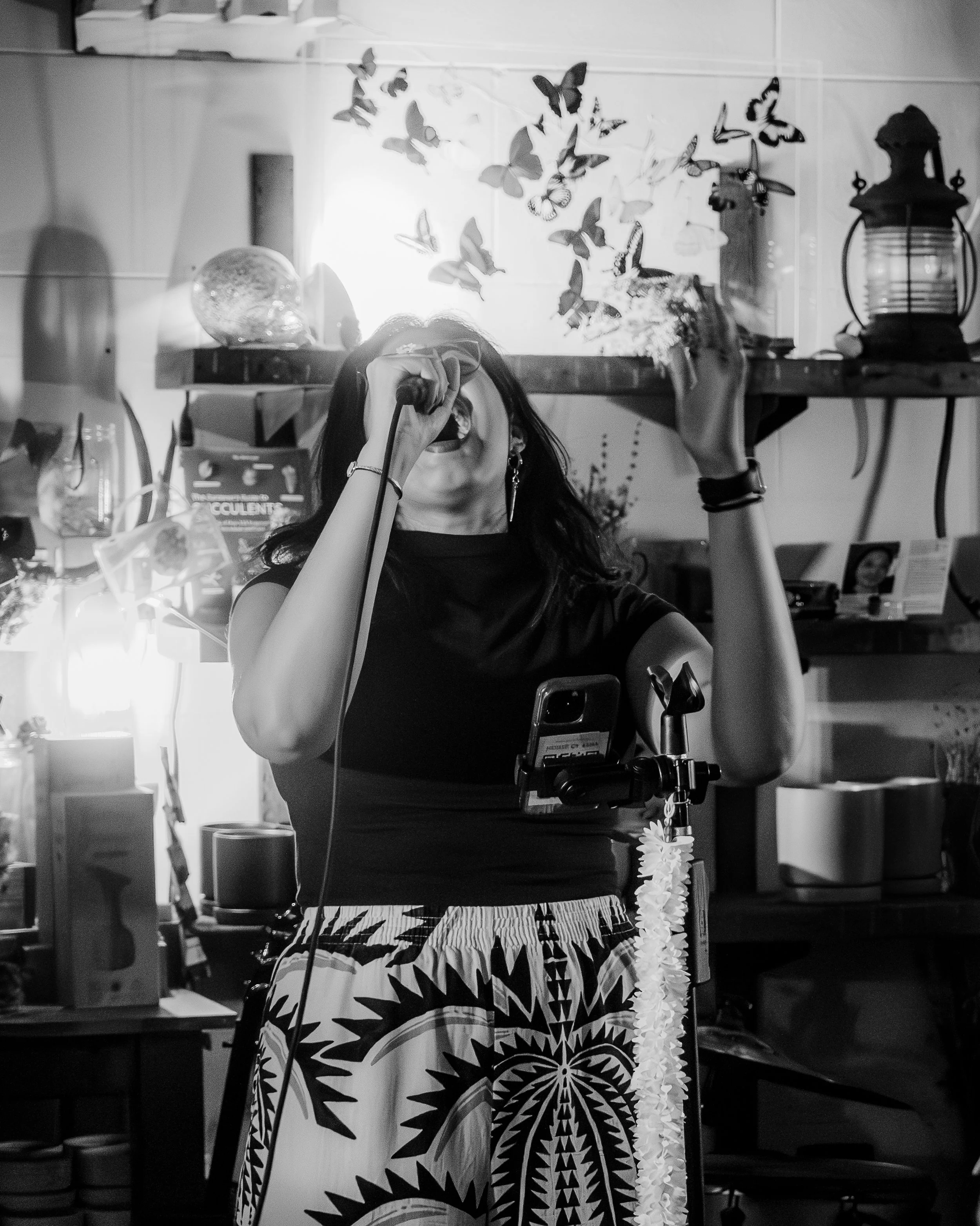 A woman singing into a microphone during a live performance in a room with various decorations like butterflies, lanterns, and plants.