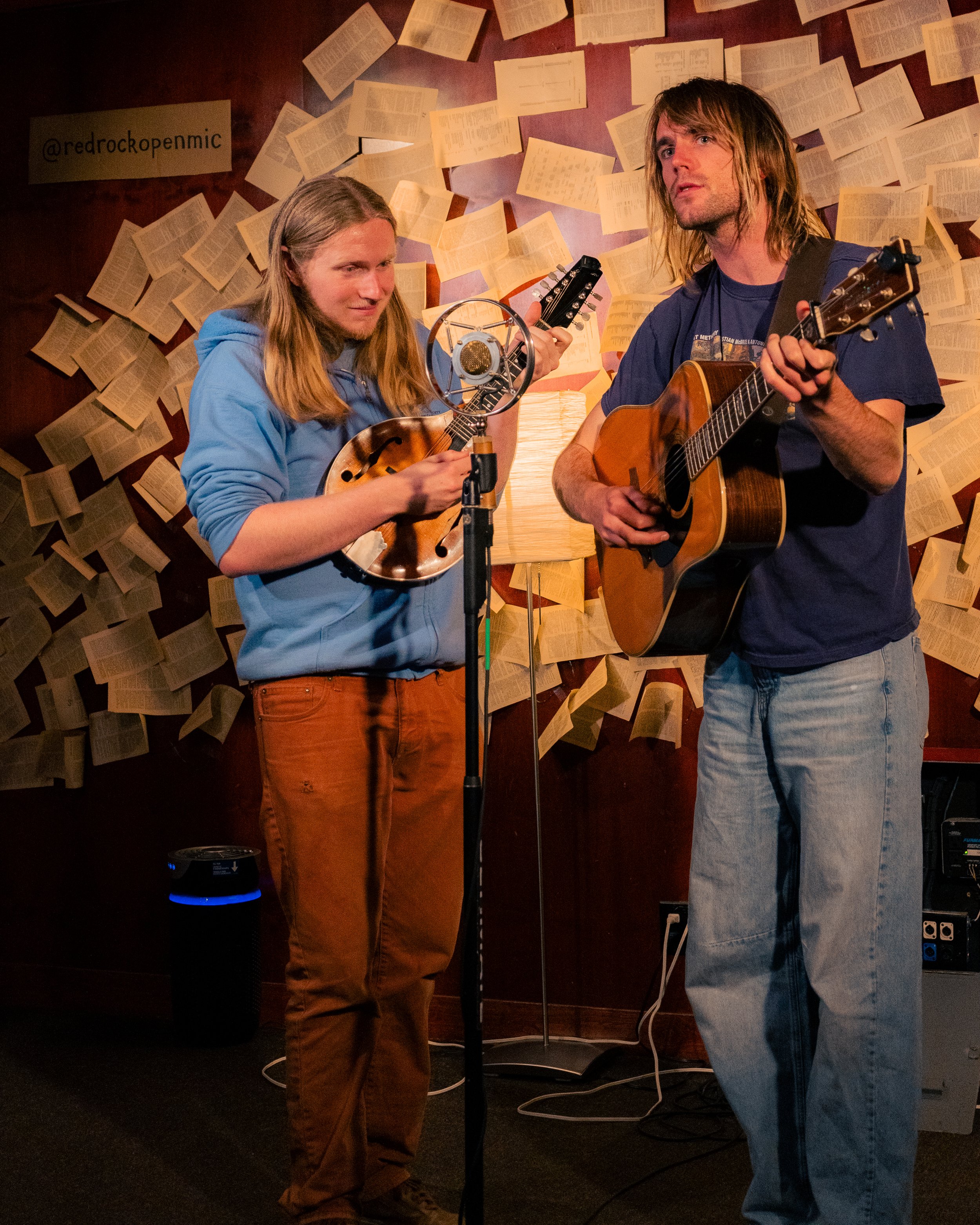 Two musicians performing with guitars and a mandolin in front of a background of floating open books.