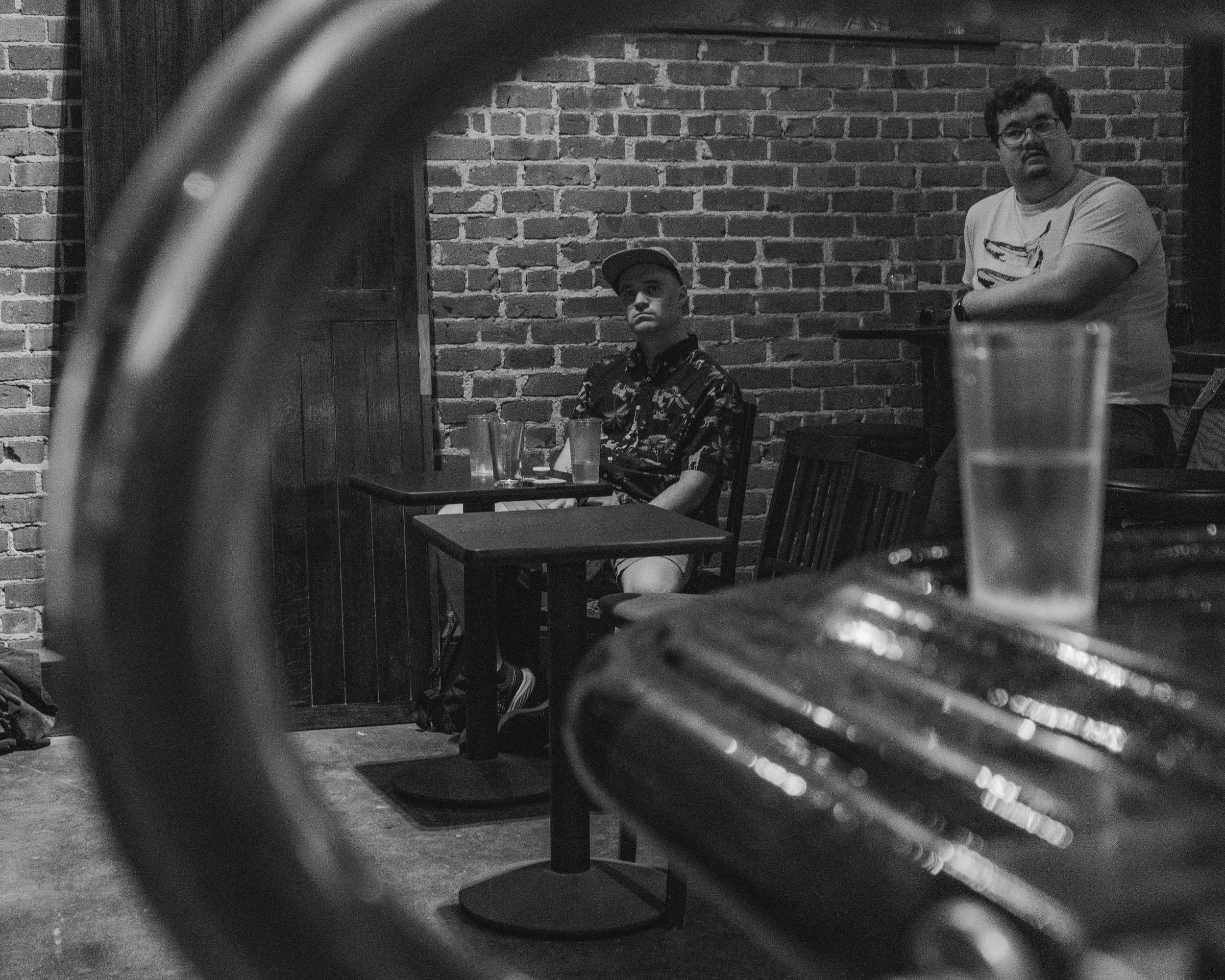 A black and white photo taken from behind a table, showing two men in a brick-walled cafe. One man is seated at a small table with drinks, wearing a cap and a patterned shirt, looking away. The other man, standing in the background, has glasses and a