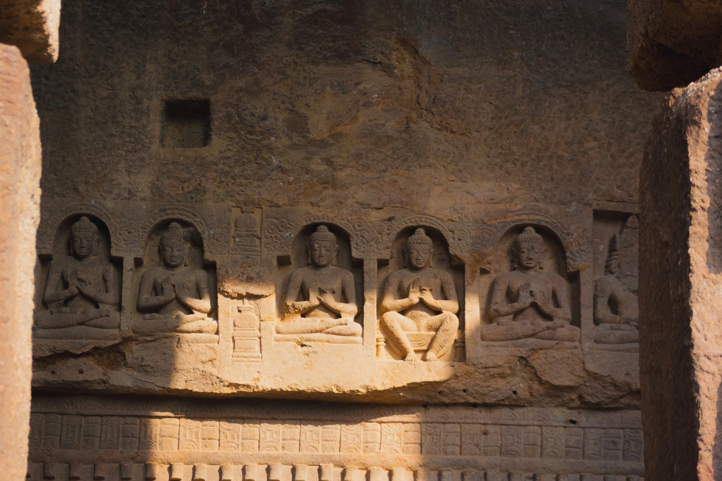 Ancient stone carvings of five seated Buddha figures in a cave, each in a meditative pose, surrounded by intricate decorative patterns.