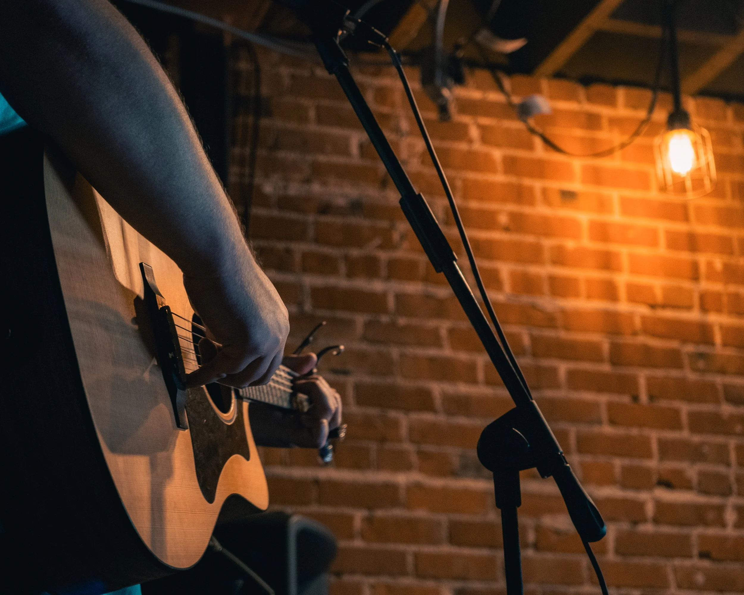 Person playing an acoustic guitar in a dimly lit room with brick walls and a hanging light bulb.