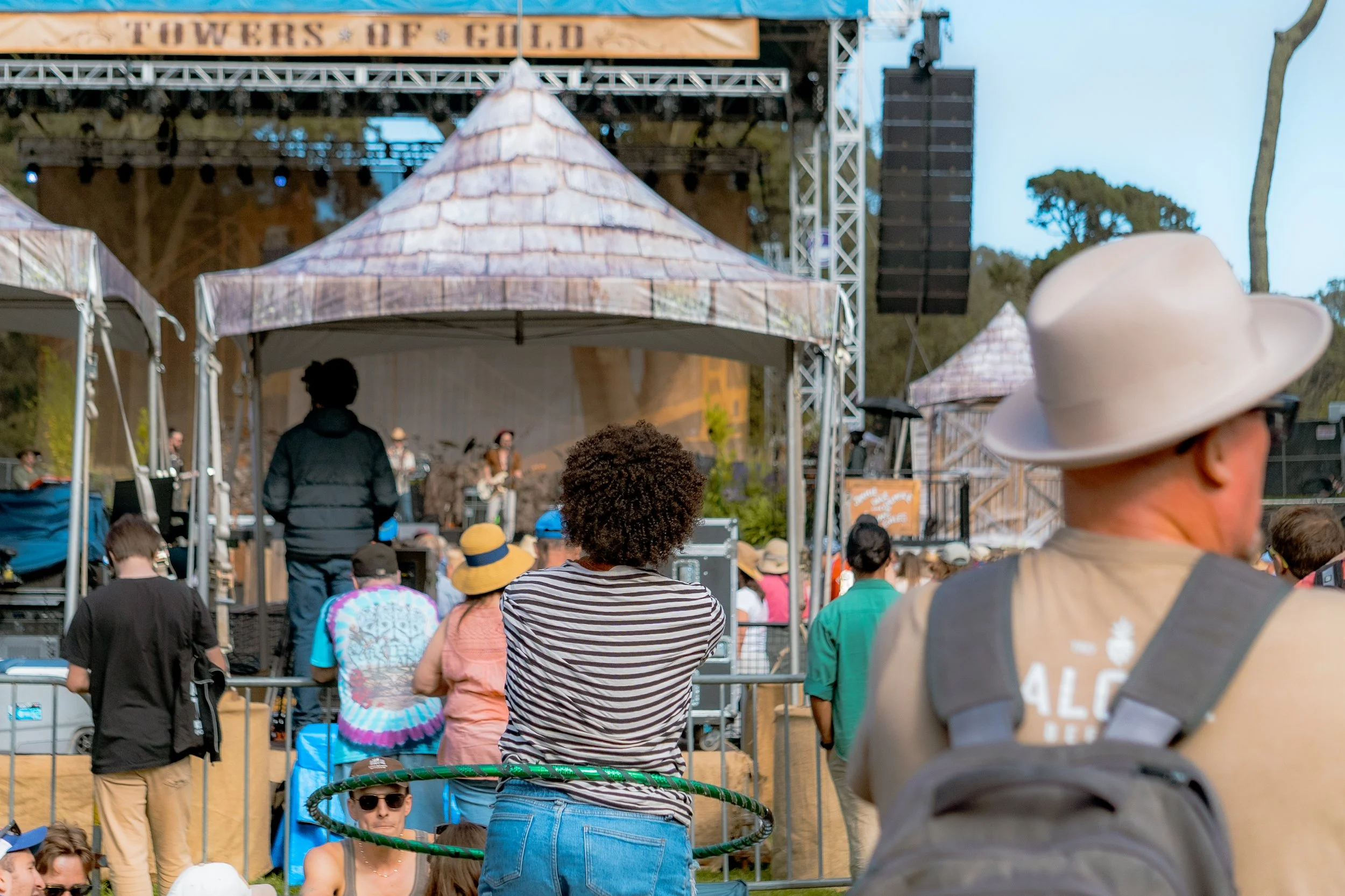 People attending an outdoor concert at a music festival, watching a band perform on stage, with some standing and others sitting, surrounded by tents and trees.