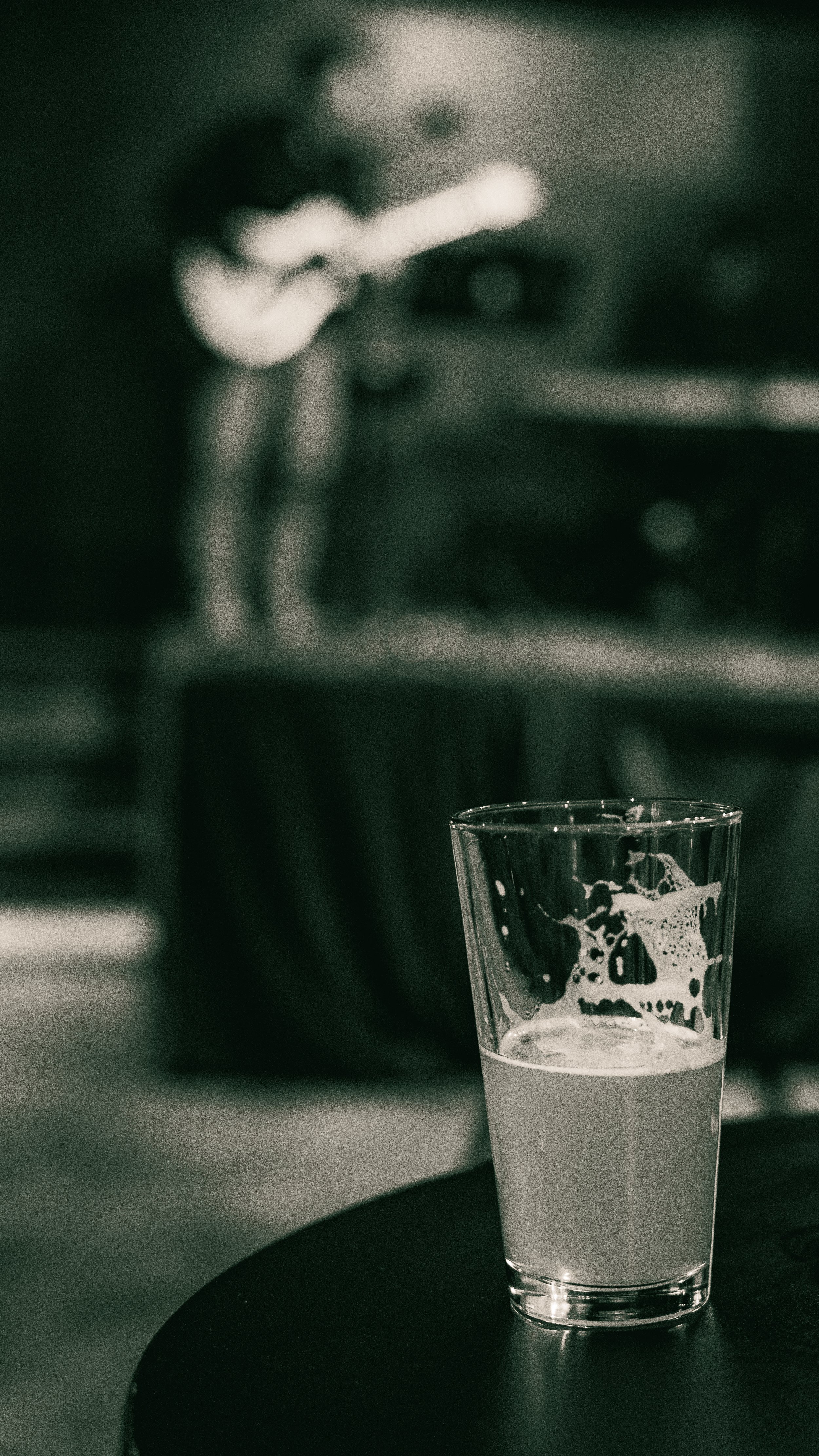 A glass partially filled with a light-colored beverage placed on a dark round table, with a blurred background of a person playing guitar.