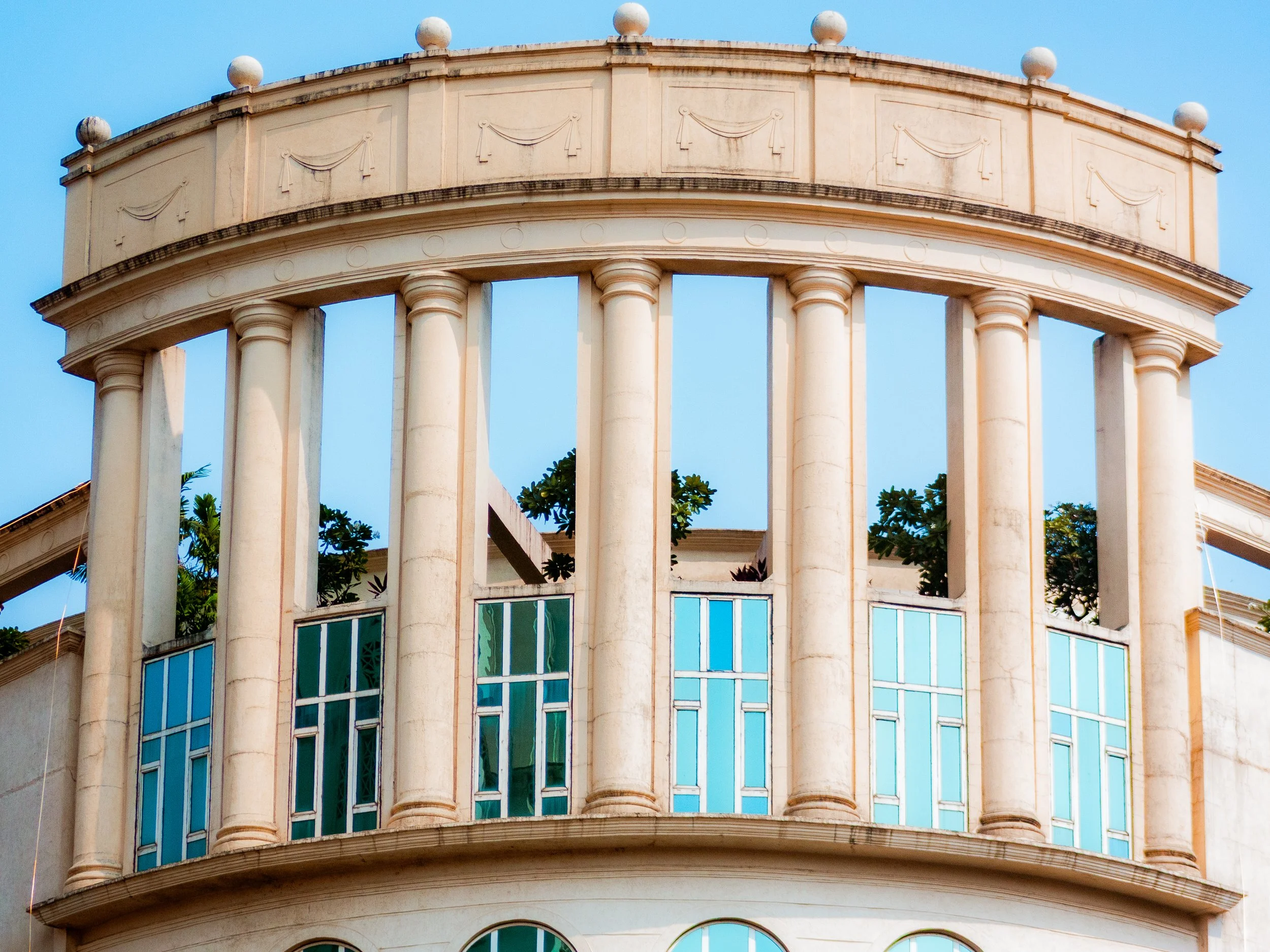A cream-colored historic building with a rounded balcony, tall columns, and large windows. The upper part has decorative panels and a row of spherical ornaments on top. Green trees are visible behind the columns.