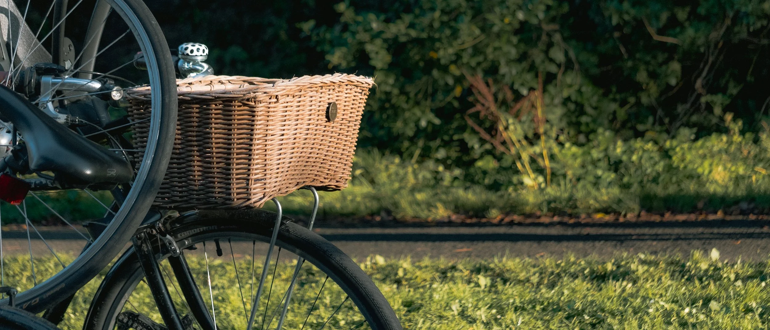 Part of a black bicycle with a wicker basket attached to the front, set against a background of green foliage and grass.
