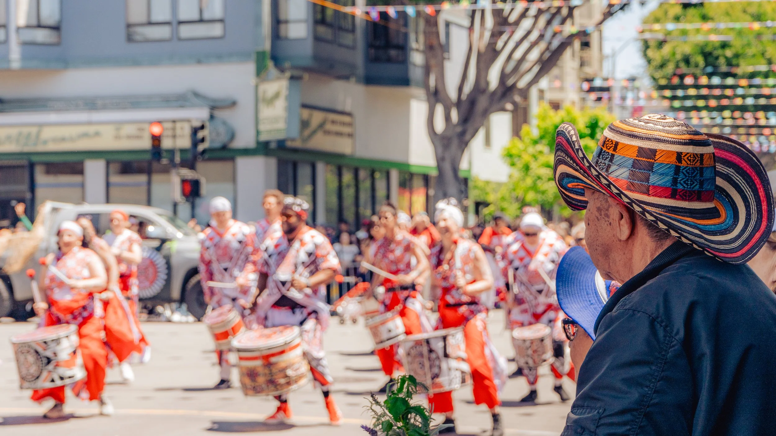 A person wearing a large, colorful, patterned sombrero, watching a parade of drummers in orange and white costumes on a city street with buildings, trees, and string lights overhead.