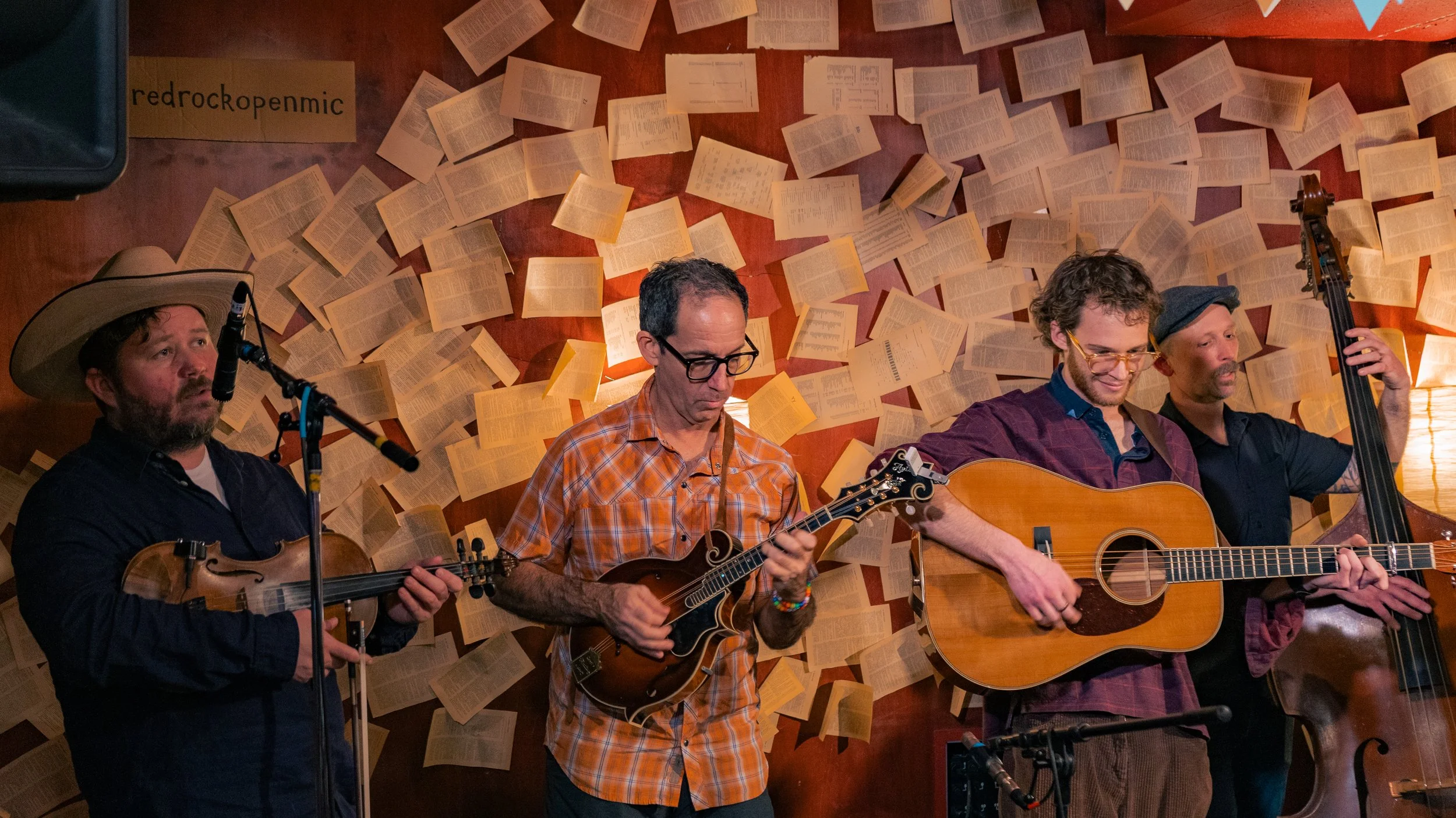 Four male musicians performing on stage with a wall of open books in the background. The first musician on the left is playing a violin and wearing a cowboy hat. The second musician is playing a mandolin and wearing glasses and an orange plaid shirt.
