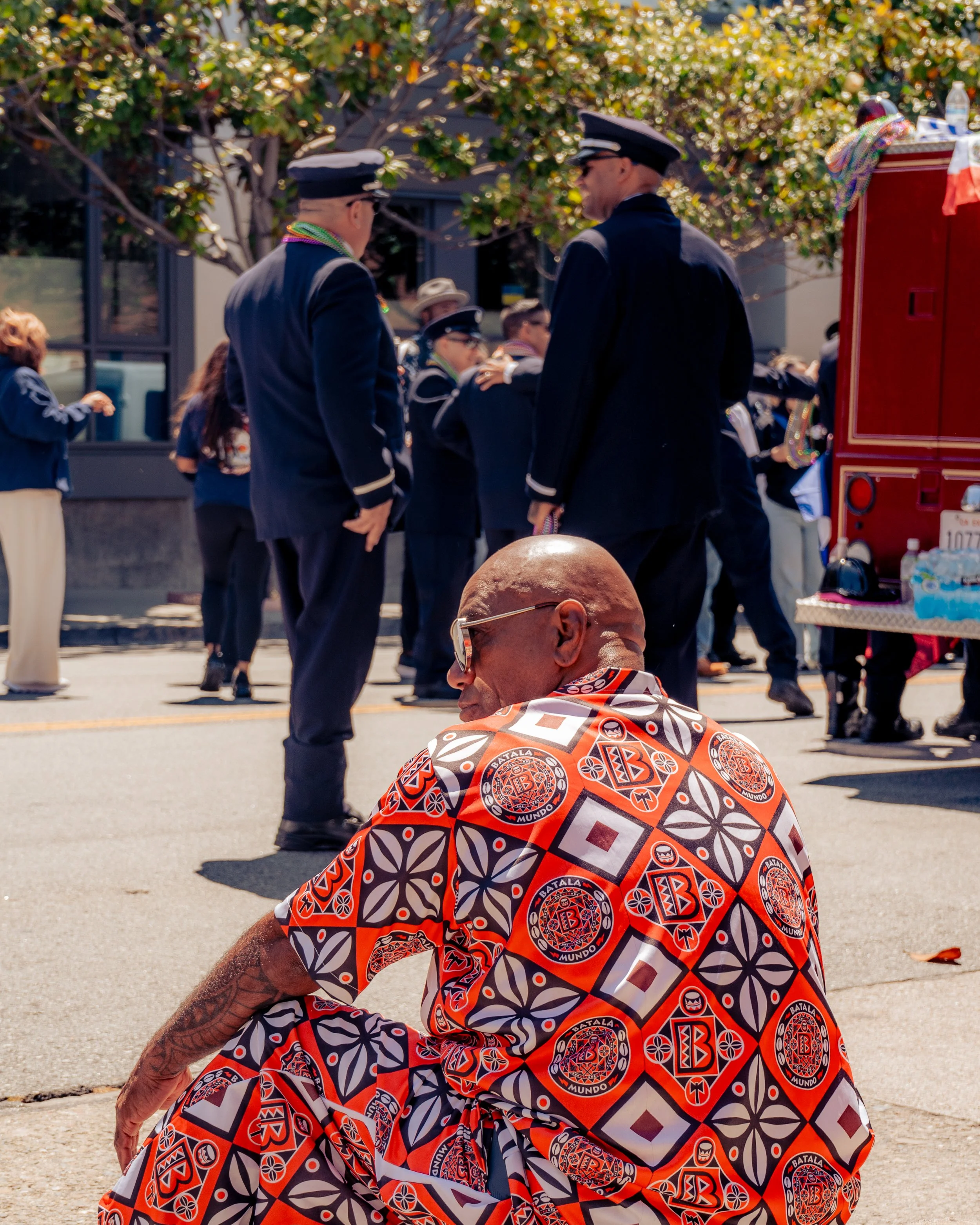 A man with glasses and a patterned shirt sitting on the sidewalk at an outdoor parade or gathering, with a group of people, some in uniform, standing and walking in the background.