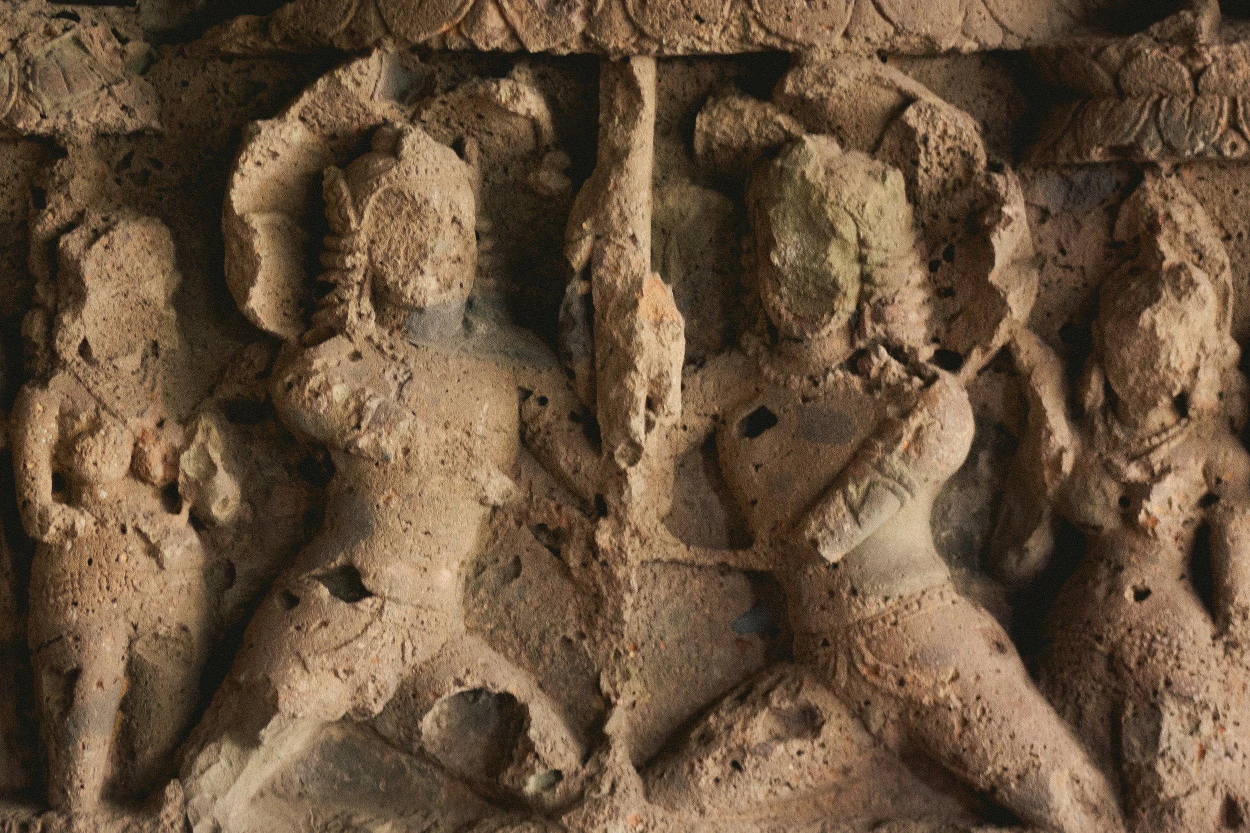 Ancient stone carving depicting figures, possibly mythological or religious, with detailed attire and headdresses, embedded in a stone wall.