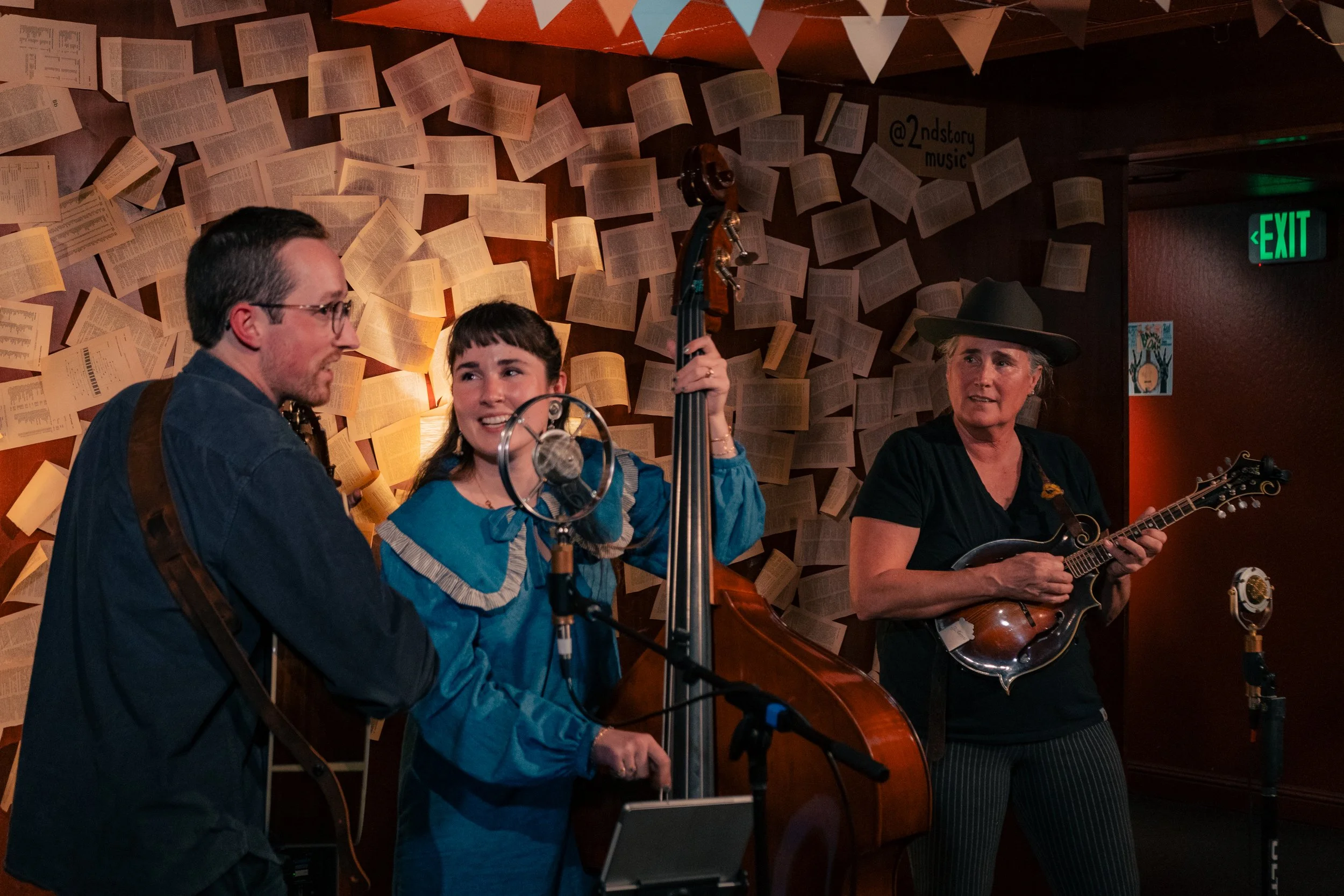 Three musicians performing on stage with a wall of open book pages behind them. One woman is playing upright bass, another woman is singing into a microphone, and a third woman is playing a mandolin. Bunting decorations are hanging overhead, and ther