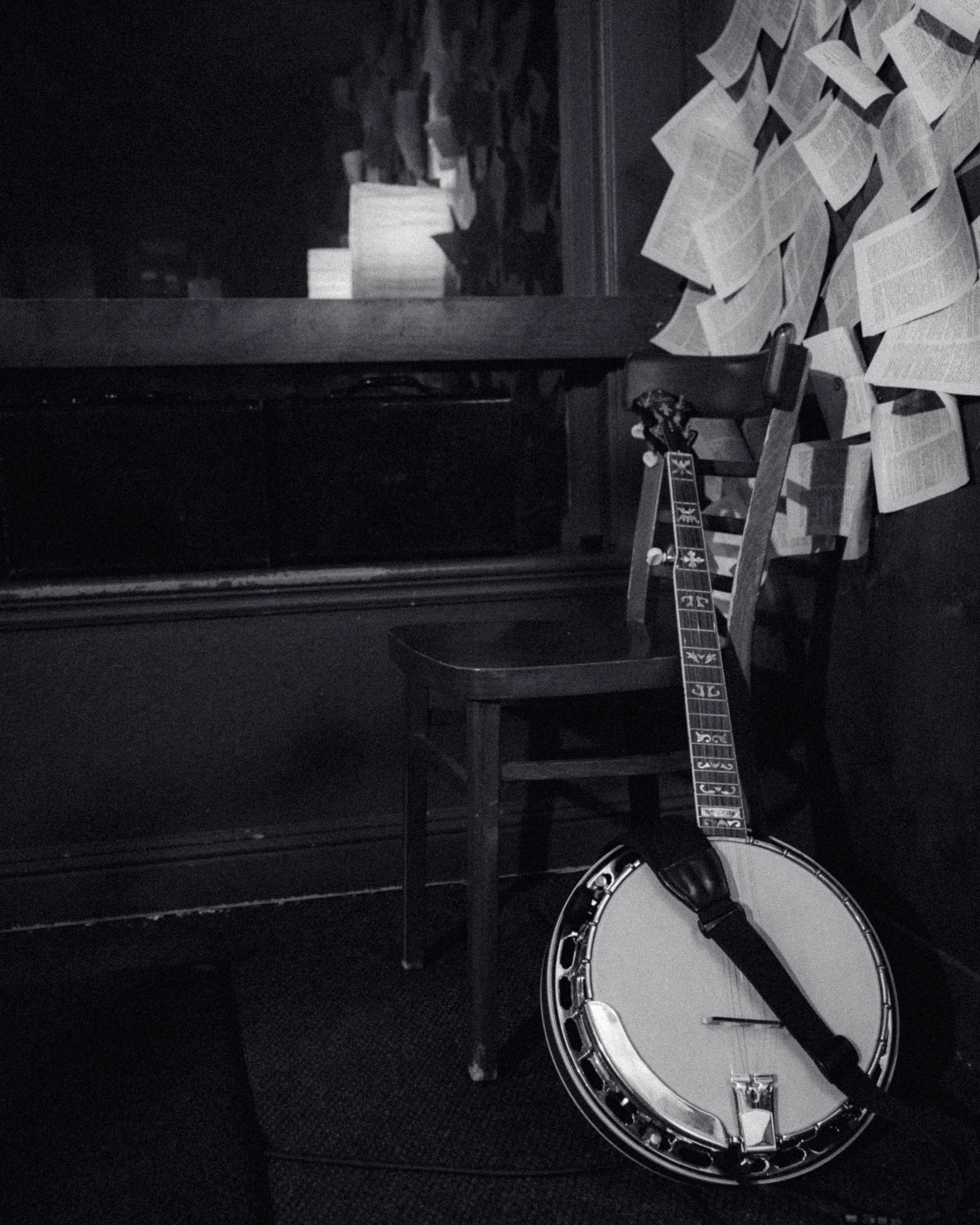 A tambourine leaning against a wooden chair in a dimly lit room, with numerous open books scattered on the floor and a window with additional books on the windowsill.