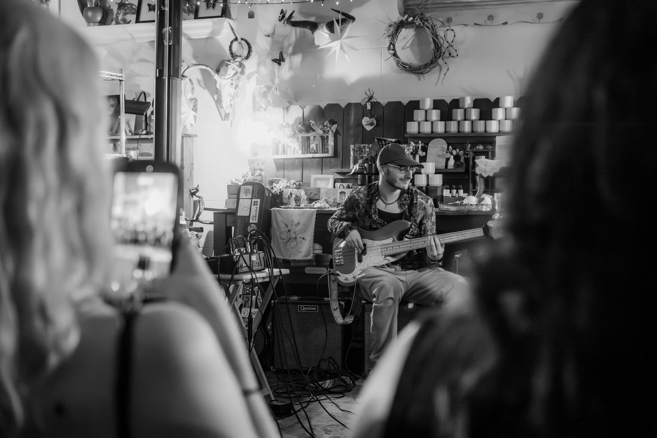 A man playing bass guitar in a cozy, decorated indoor space with two women in the foreground, one recording the performance on a smartphone. The background includes shelves with candles, decorative items, and hanging ornaments.