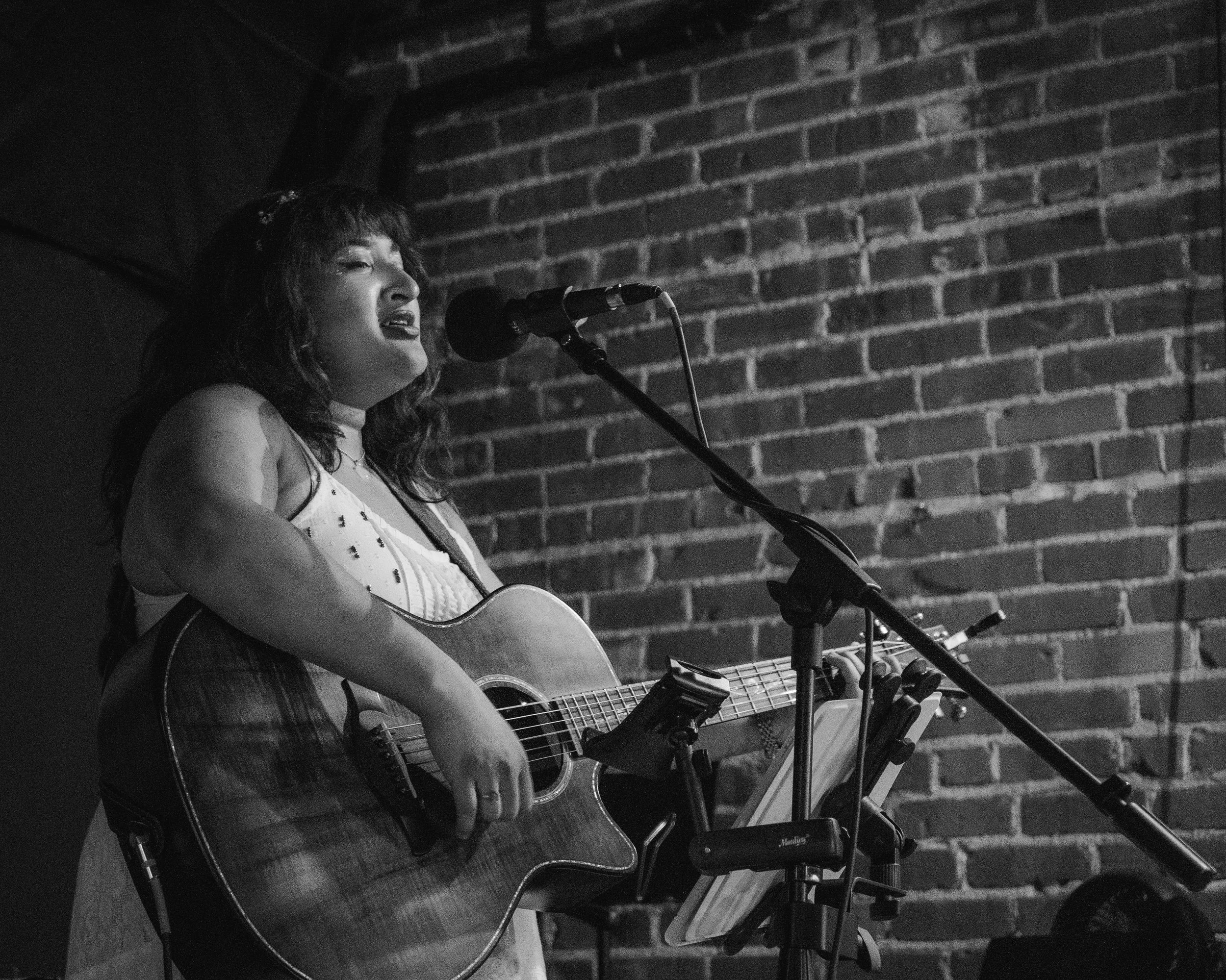 A woman with dark hair singing and playing an acoustic guitar on stage, with a brick wall background.