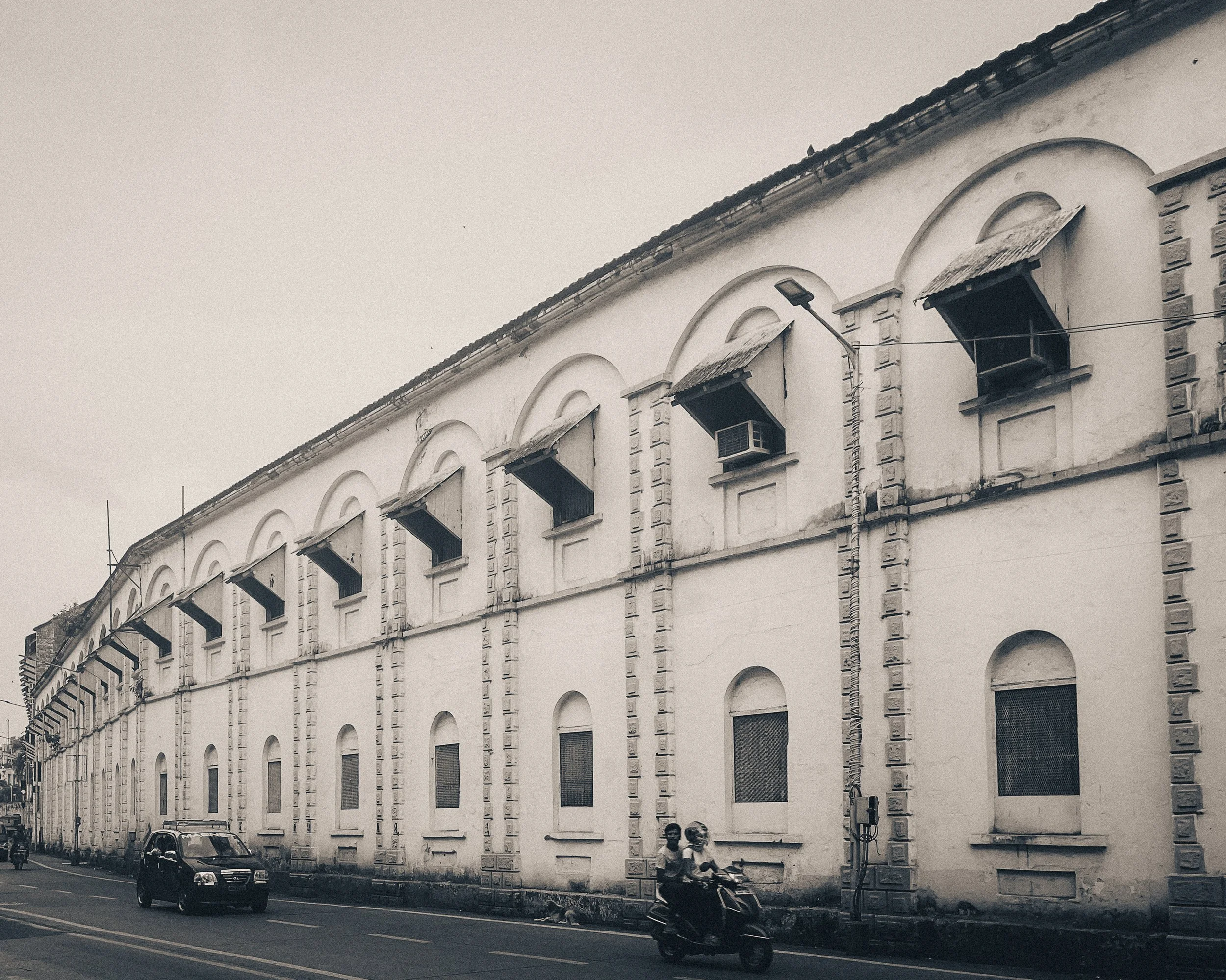 A black and white photo of a long, old building with arched windows and air conditioning units on the upper windows. Two people are riding a motorcycle on the street in front of the building, and a car is driving by. There is a streetlight and electr