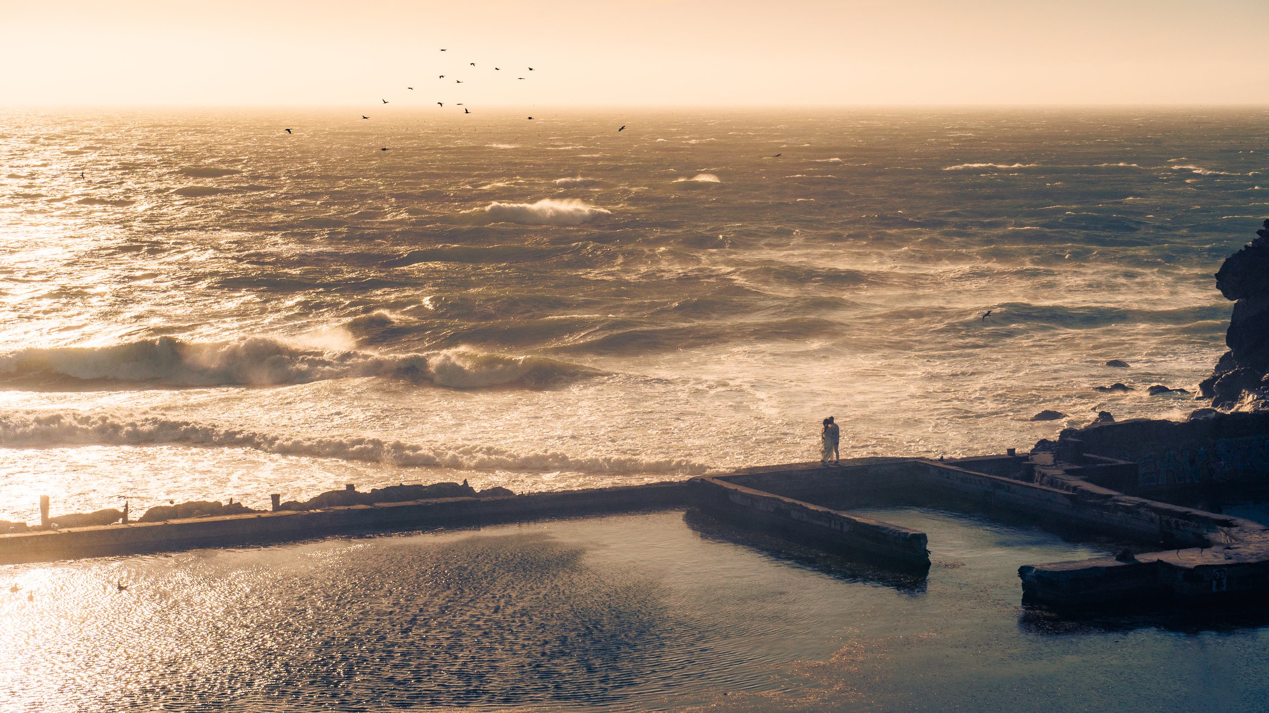 A person standing near the edge of a seaside swimming pool during sunset, with waves crashing in the ocean and birds flying overhead.
