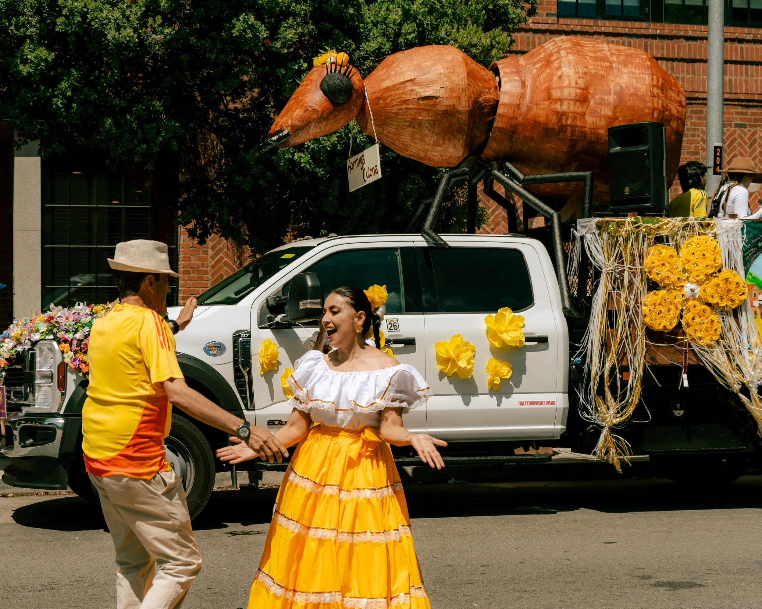 People dancing in front of a parade float decorated with yellow flowers and a large ant sculpture on top.