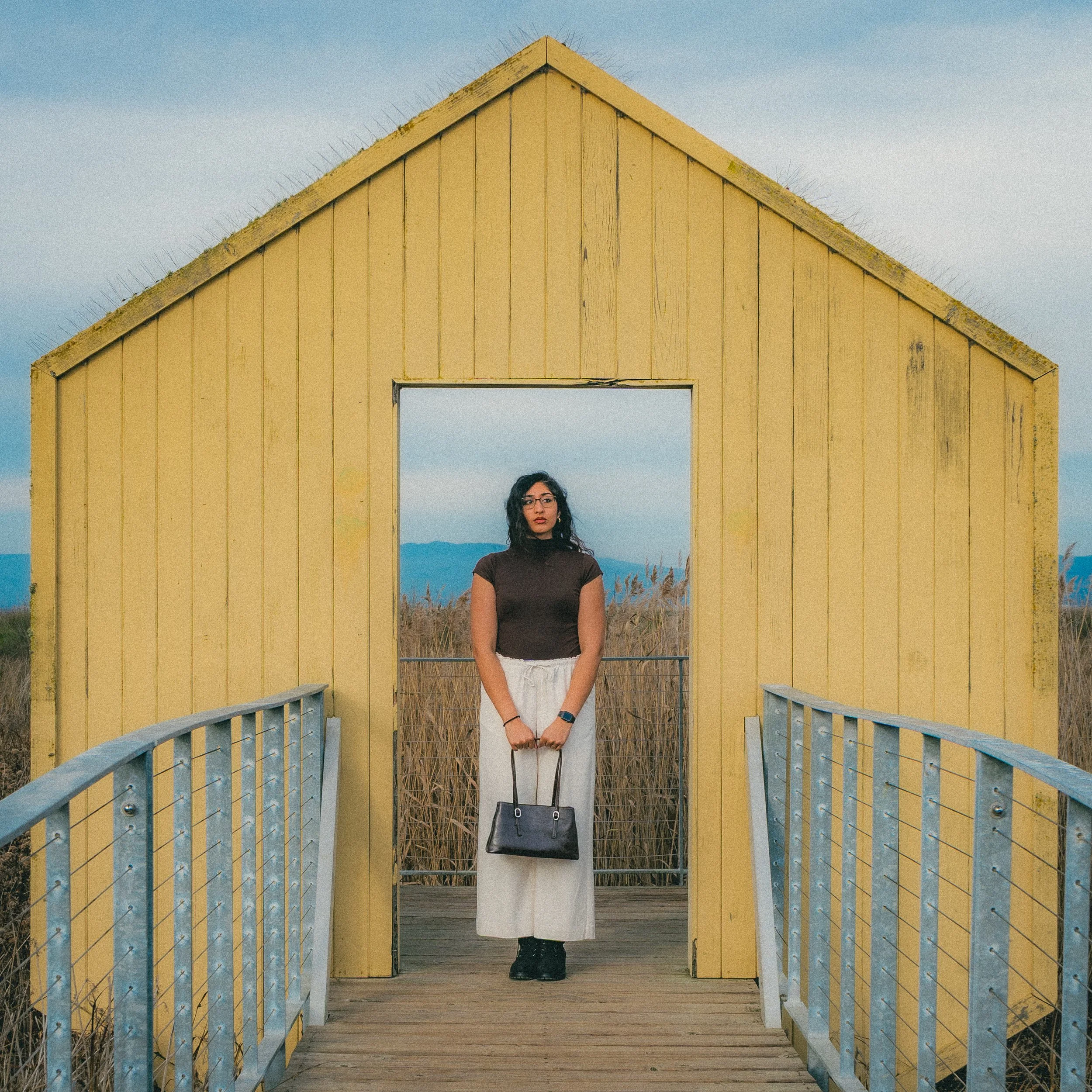 A woman standing on a wooden bridge in front of a yellow wooden hut with an open doorway, surrounded by tall grass, with mountains in the background under a cloudy sky.