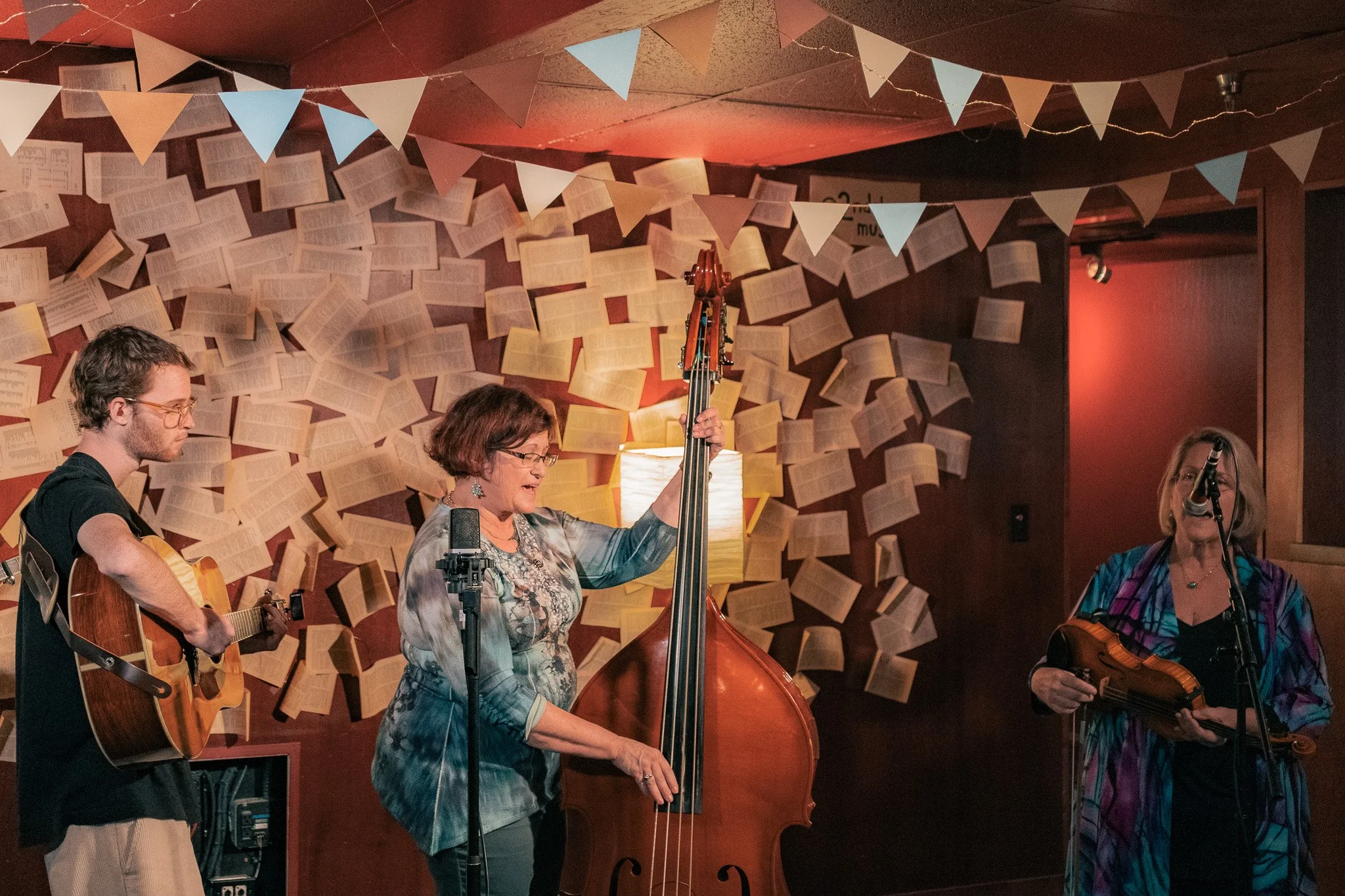Three musicians performing with guitars and a double bass in a decorated indoor setting with open books on the wall and triangular flags hanging from the ceiling.