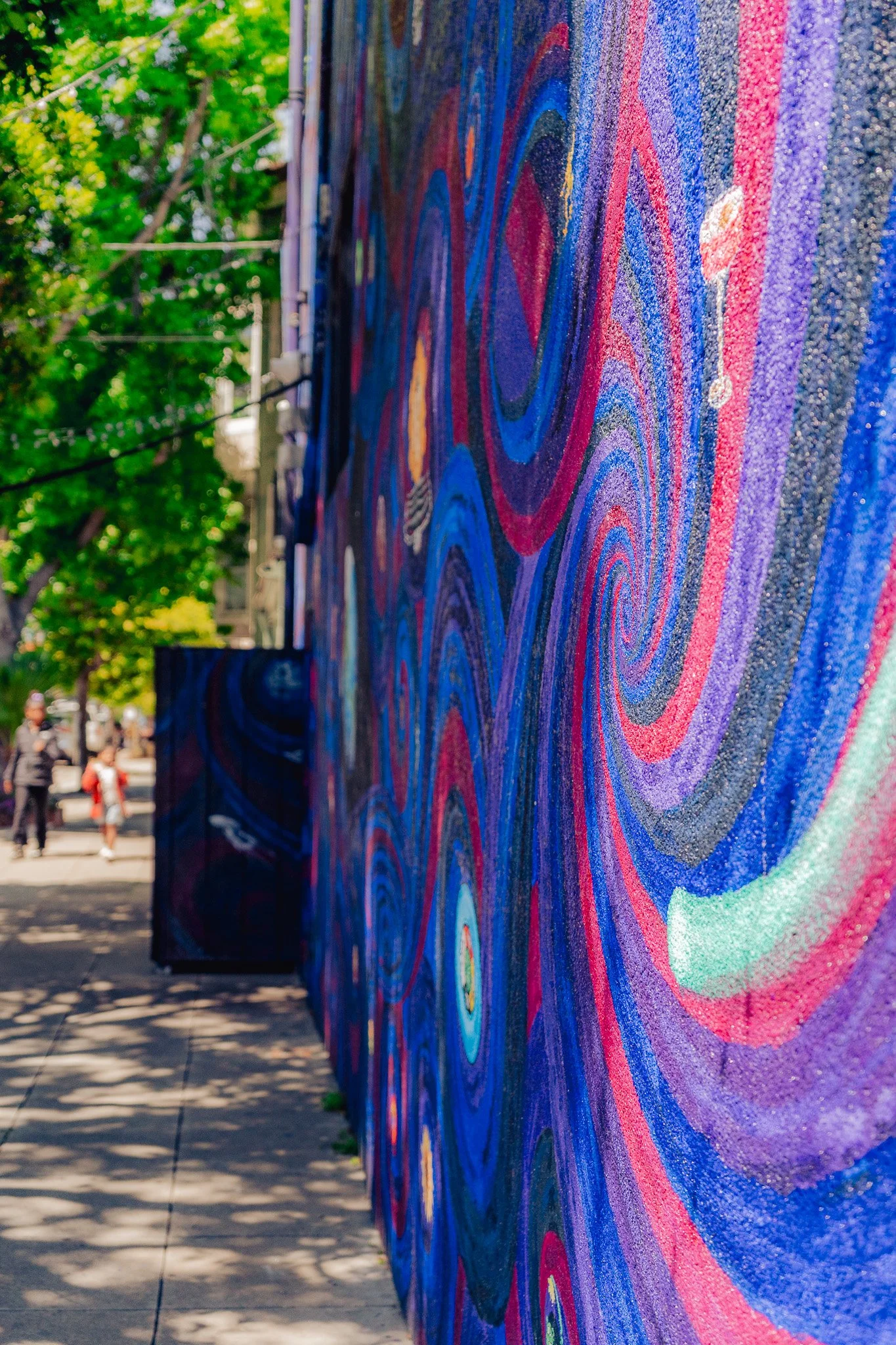 Close-up of a large colorful abstract mural with swirling patterns in red, blue, purple, and green on an outdoor wall.