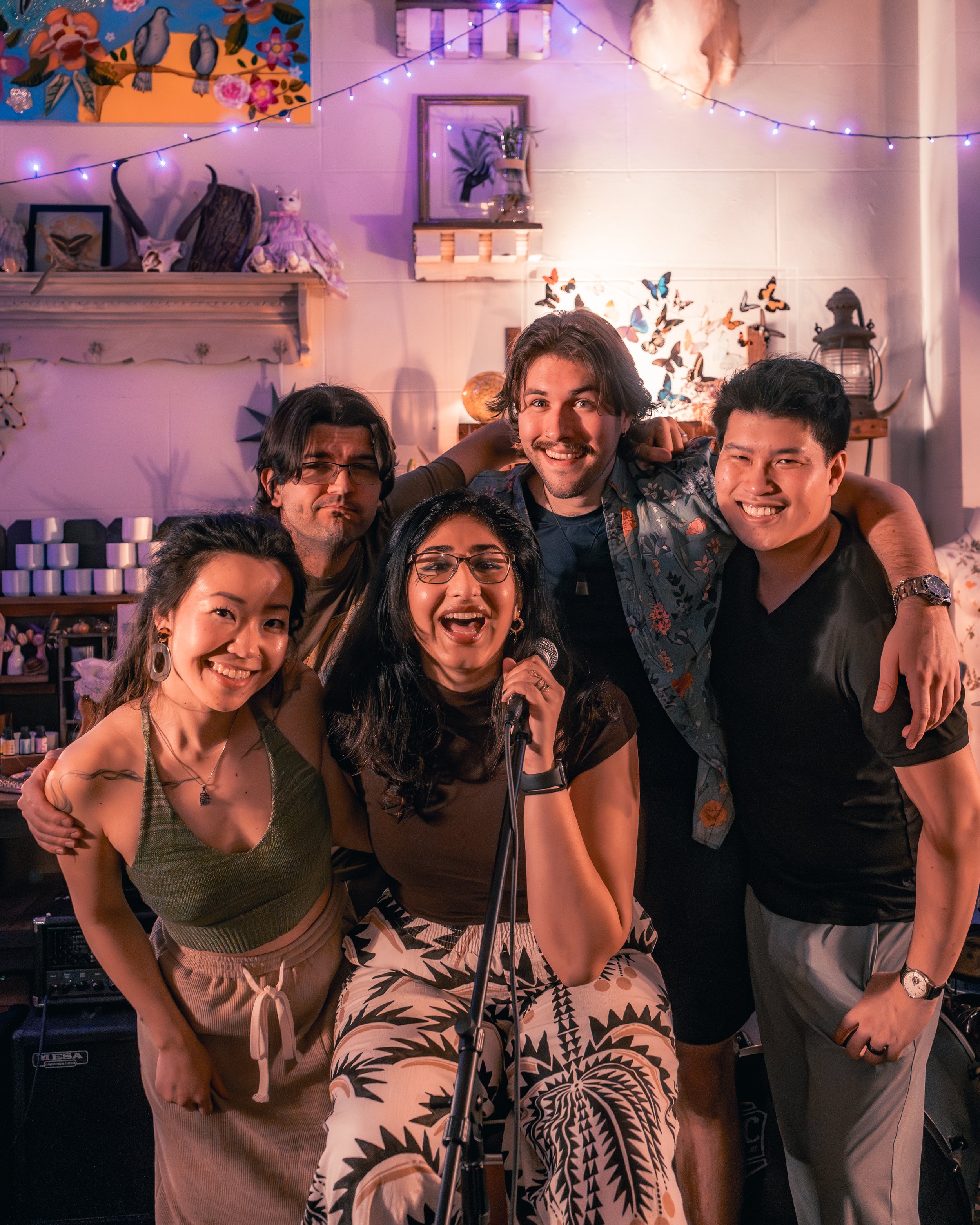 A group of five friends smiling and posing together at a party or gathering, with a woman holding a microphone. The background features colorful decorations, string lights, and artwork, creating a lively and festive atmosphere.