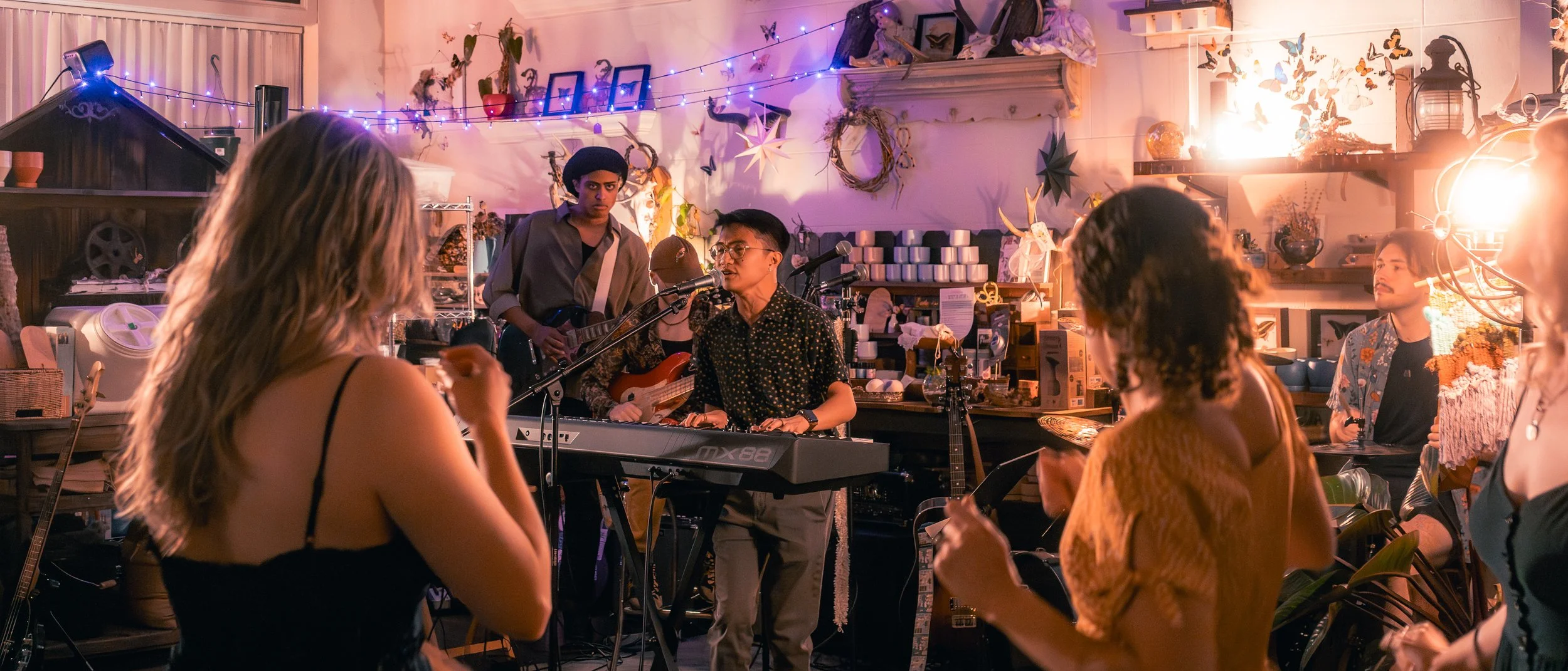 A group of musicians performing live in a cozy, decorated indoor venue. The scene includes a keyboard player, a singer, a guitarist, and several audience members enjoying the music. Colorful string lights, artwork, and decorative items adorn the back