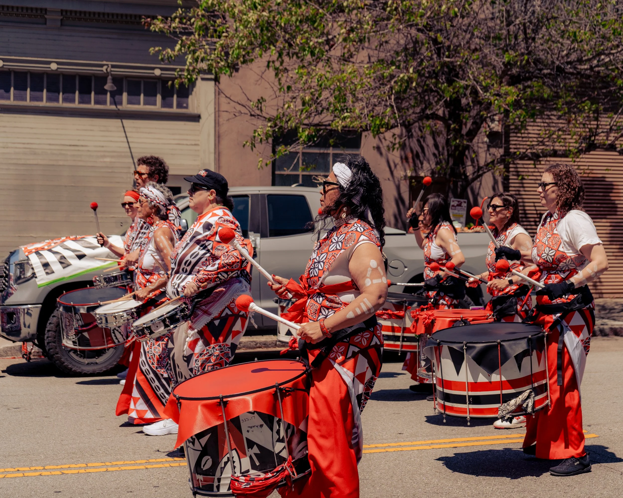 A group of women participating in a parade, playing drums and percussion instruments, wearing coordinated red, black, and white patterned outfits, walking along a street with buildings and parked cars in the background.