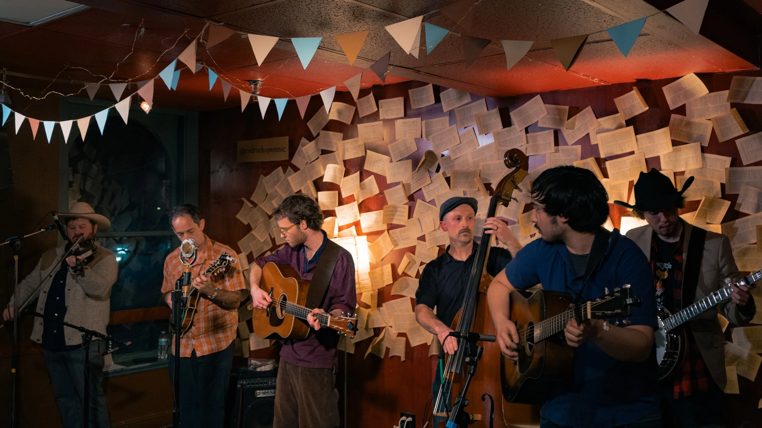 Group of six musicians performing on stage with books and bunting decorations behind them, playing guitars, a double bass, and singing into microphones, in a cozy, dimly lit venue.
