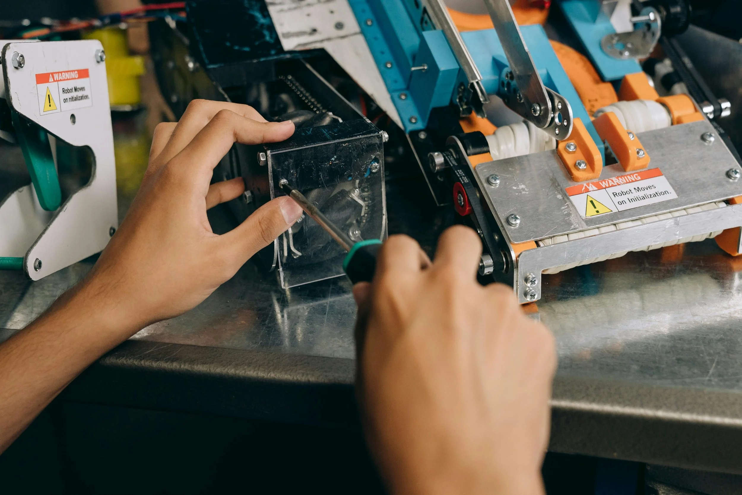 Person working on a mechanical and electronic device with a screwdriver, with warning labels that say "Warning: Robot Moves on Initialization."
