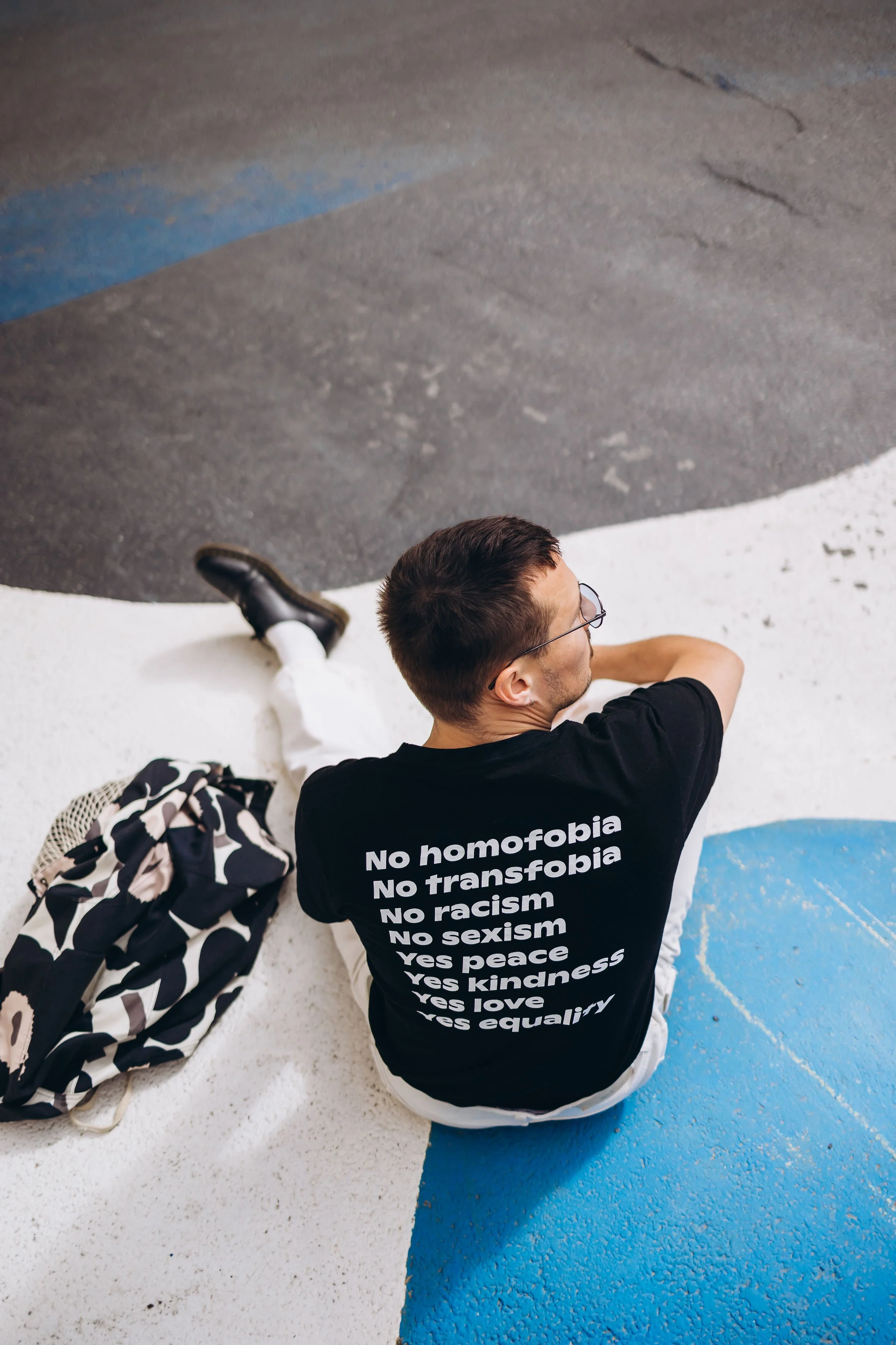 Pesojoonas istuu maassa, katsoo toiseen suuntaan, päällä musta t-paita, jossa on tekstiä, ja on vieressä vaalea ja musta kuviollinen reppu.