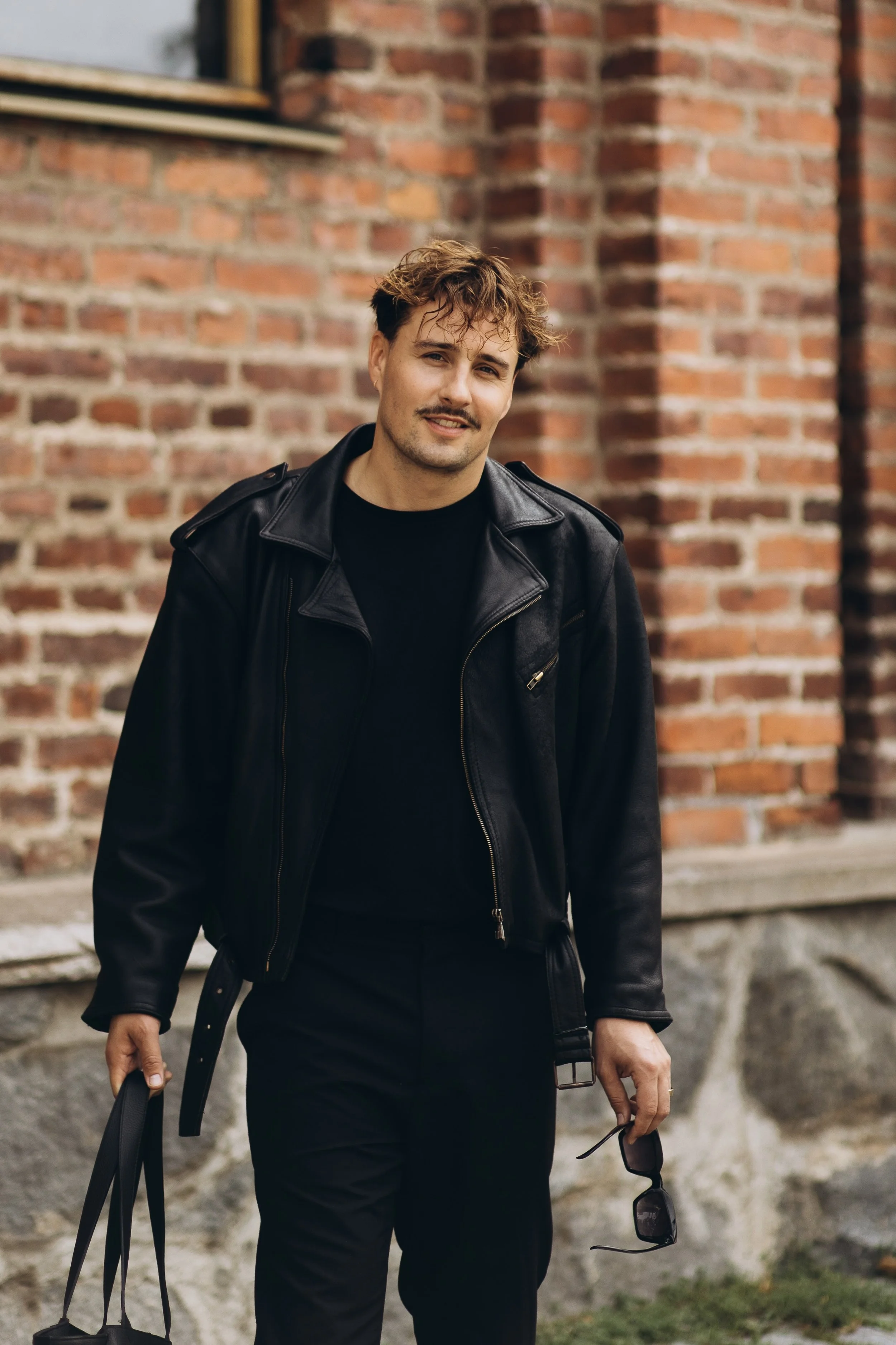 kävelee kaupungin katukäytävällä, pukeutuneena mustaan nahkatakkiin ja mustiin housuihin, ja hän kantaa käsilaukkuja ja aurinkolaseja.